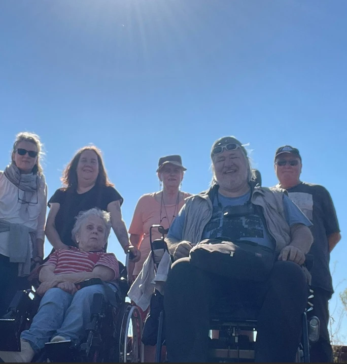 Gruppe von sechs Personen, darunter zwei im Rollstuhl, vor hellem Himmel mit Sonnenstrahlen