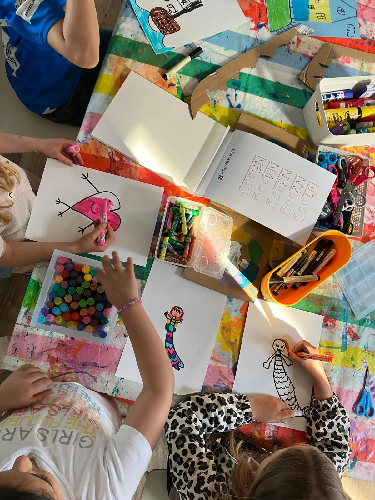 Kinder sitzen an einem bunt gedeckten Tisch und malen bunte Bilder mit Stiften und Filzstiften