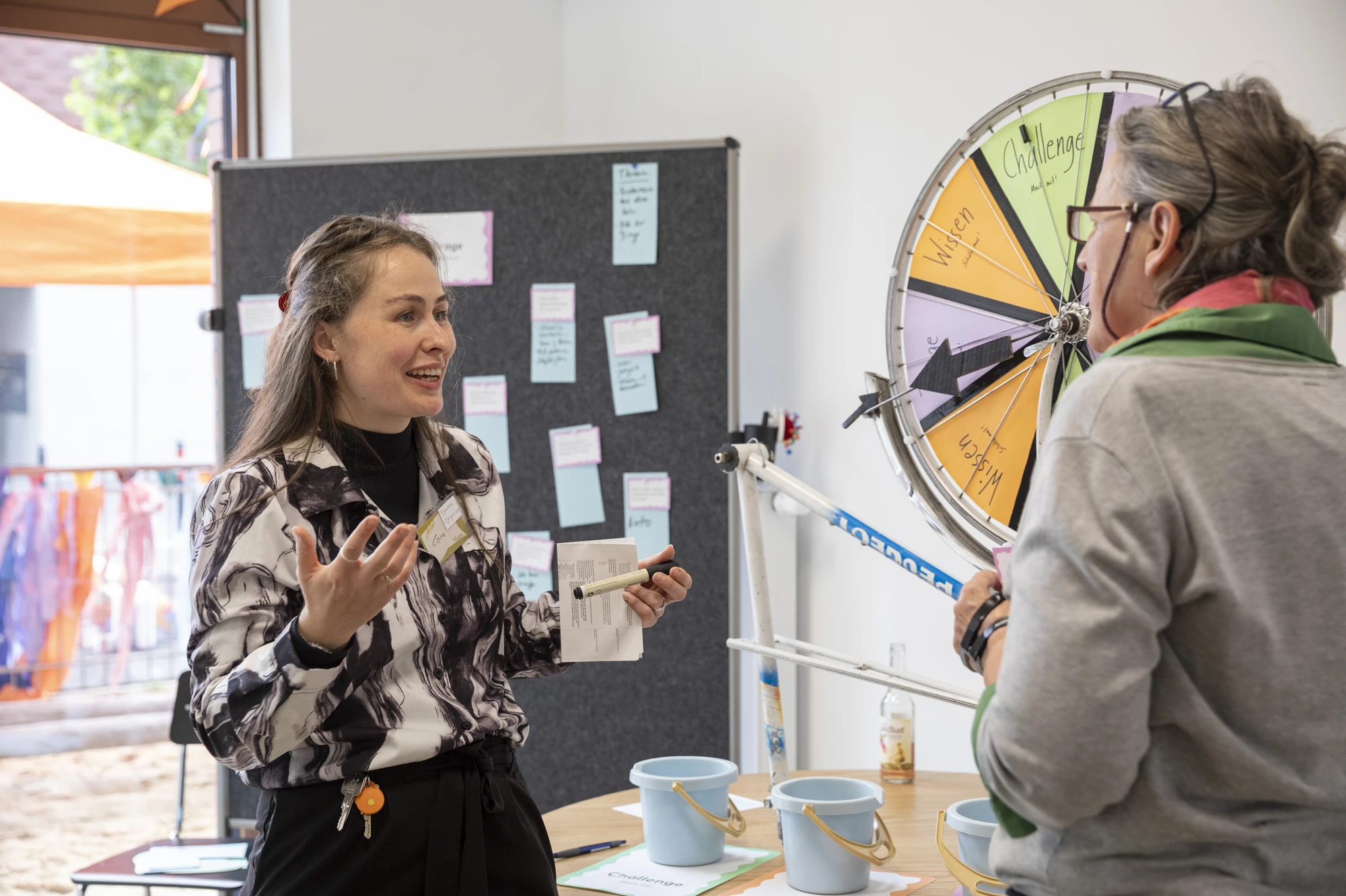 Cora Sabisch steht vor einer Pinnwand und spricht gestikulierend zu einer Frau, die nur von hinten zu sehen ist. Auf einem Tisch stehen kleine Eimer und darauf ein Gl&uuml;cksrad.