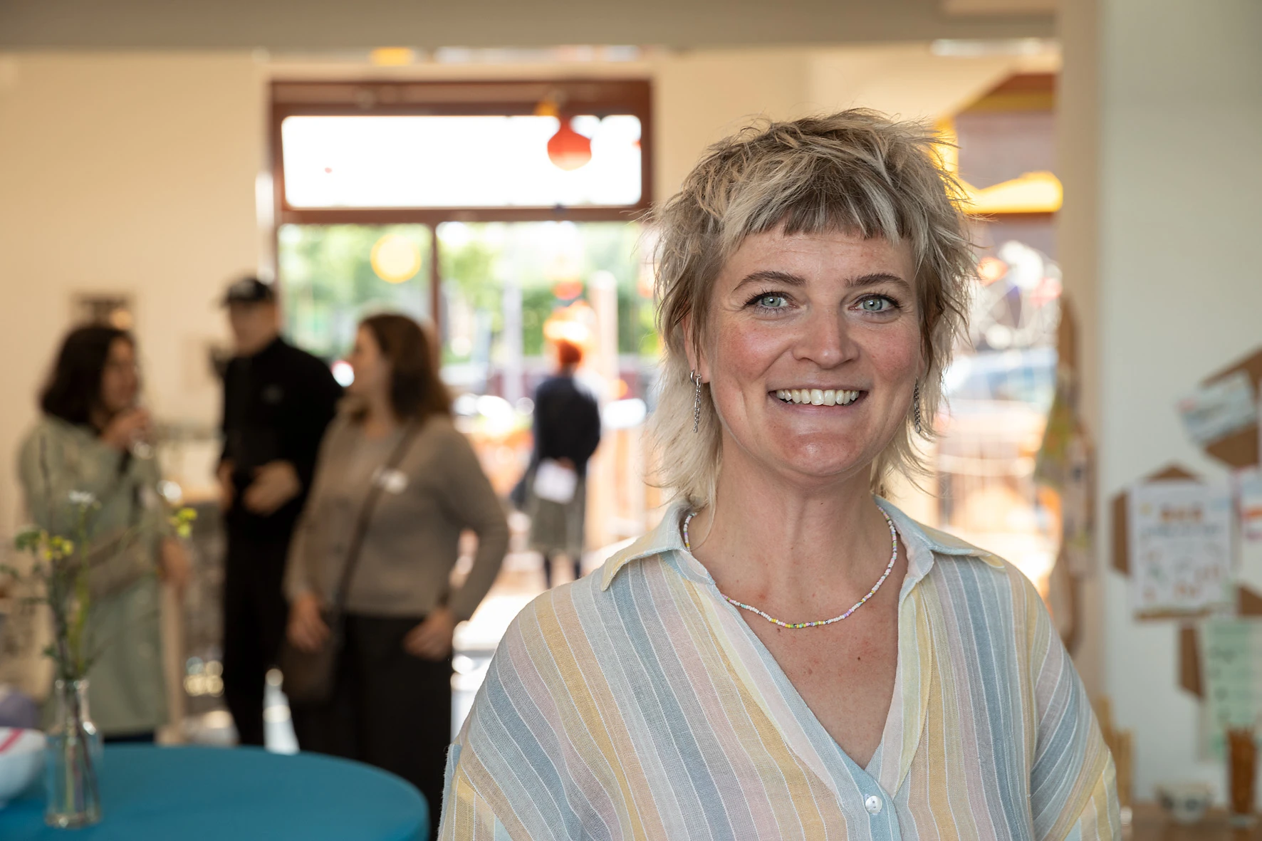Frau mit kurz geschnittenem Haar und gestreiftem Hemd steht in einem hellen Raum mit mehreren Personen im Hintergrund.