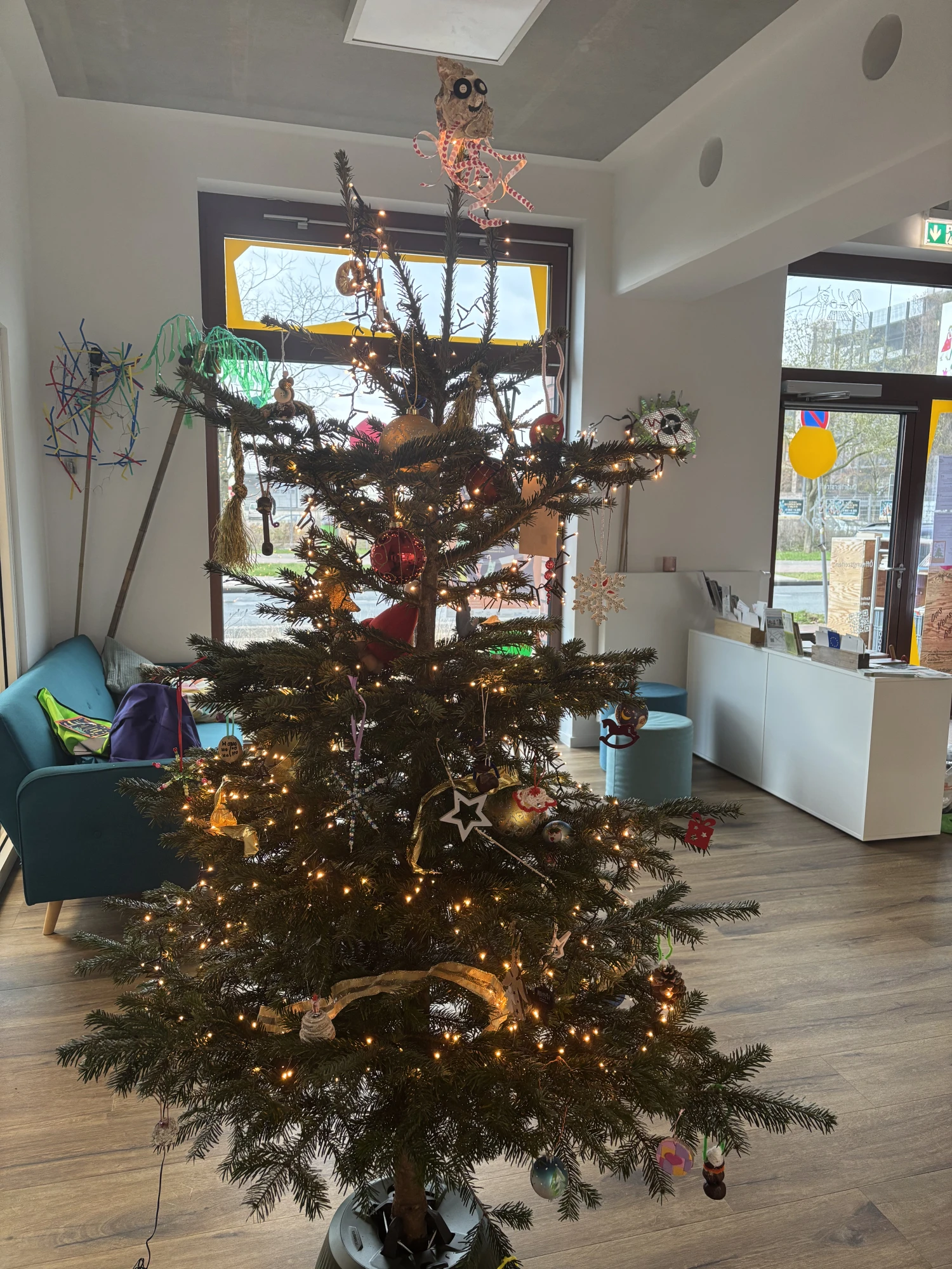 Ein mittelgro&szlig;er, geschm&uuml;ckter Tannenbaum steht in einem hellen Raum mit gro&szlig;en Fenstern. Am Baum h&auml;ngen verschiedene bunte, selbstgemachte Weihnachtsanh&auml;nger, darunter Kugeln, Sterne, kleine Figuren und Bastelarbeiten. Auf der Baumspitze sitzt eine humorvolle, gebastelte Figur mit gro&szlig;en Augen. Der Baum ist mit warmwei&szlig;en Lichterketten beleuchtet. Im Hintergrund stehen ein t&uuml;rkisfarbenes Sofa mit Taschen darauf, bunte Dekorationen an den W&auml;nden und eine wei&szlig;e Empfangstheke. Die Atmosph&auml;re wirkt freundlich und einladend.