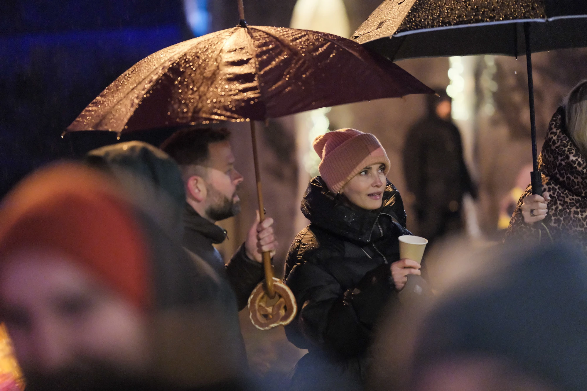 Mehrere Personen stehen im Regen, zwei halten Regenschirme, eine Person trägt eine rosa Mütze und hält einen Pappbecher.