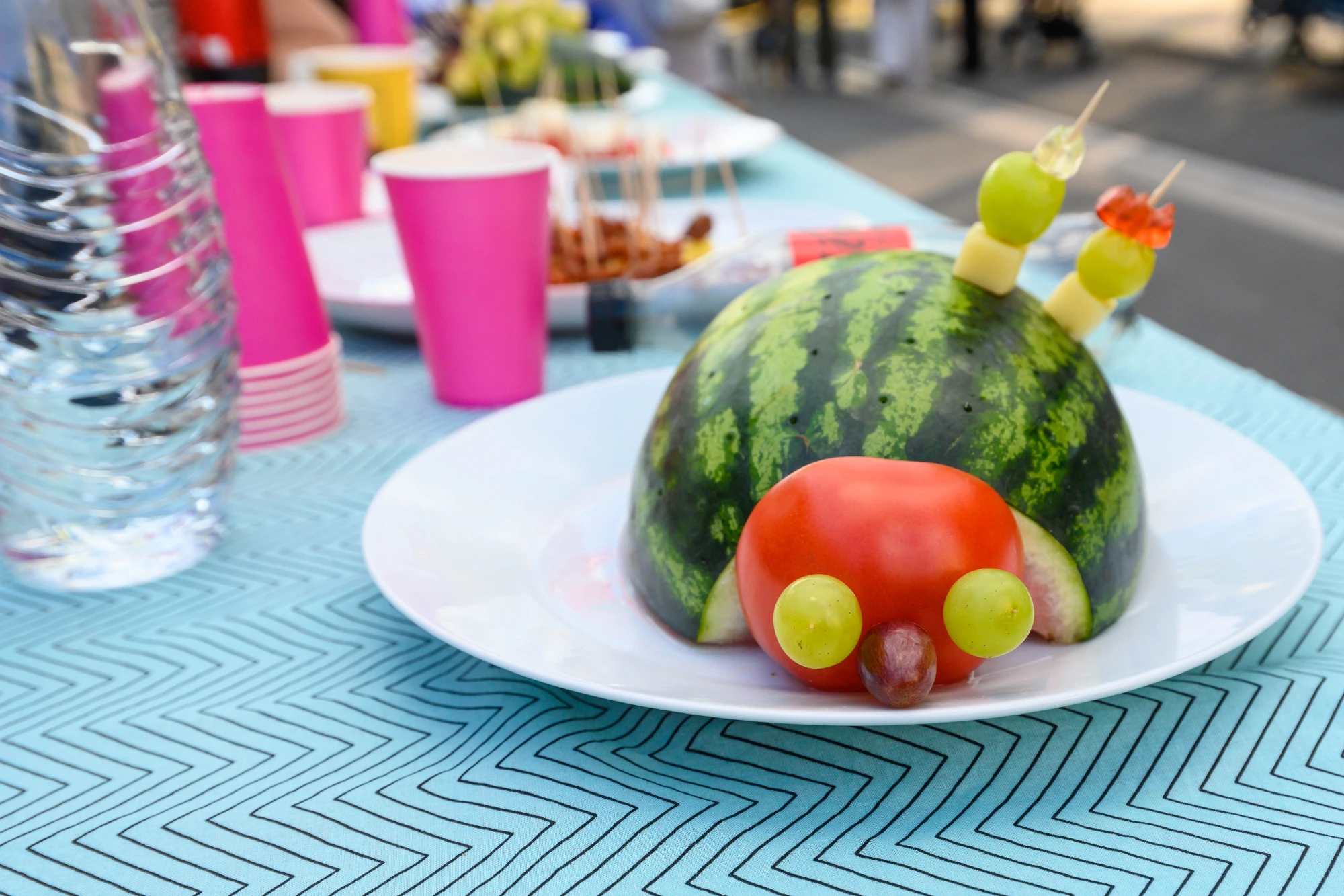 Halbe Wassermelone auf wei&szlig;em Teller, dekoriert als Schnecke mit Tomatenkopf, Traubenaugen und Spie&szlig;en als F&uuml;hler auf blau gemustertem Tisch