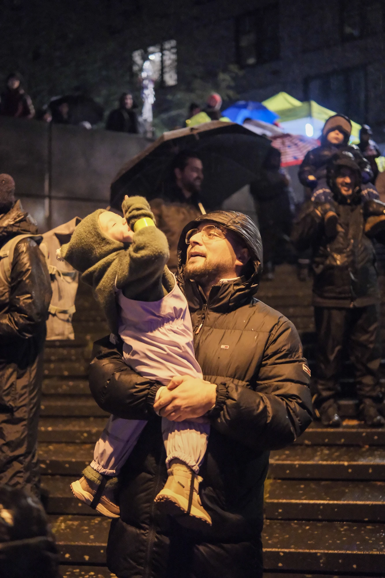 Person in dunkler Winterjacke hält Kind in lila Schneeanzug und gelber Mütze auf einer Treppe bei Nacht, im Hintergrund weitere Personen und bunte Zelte.