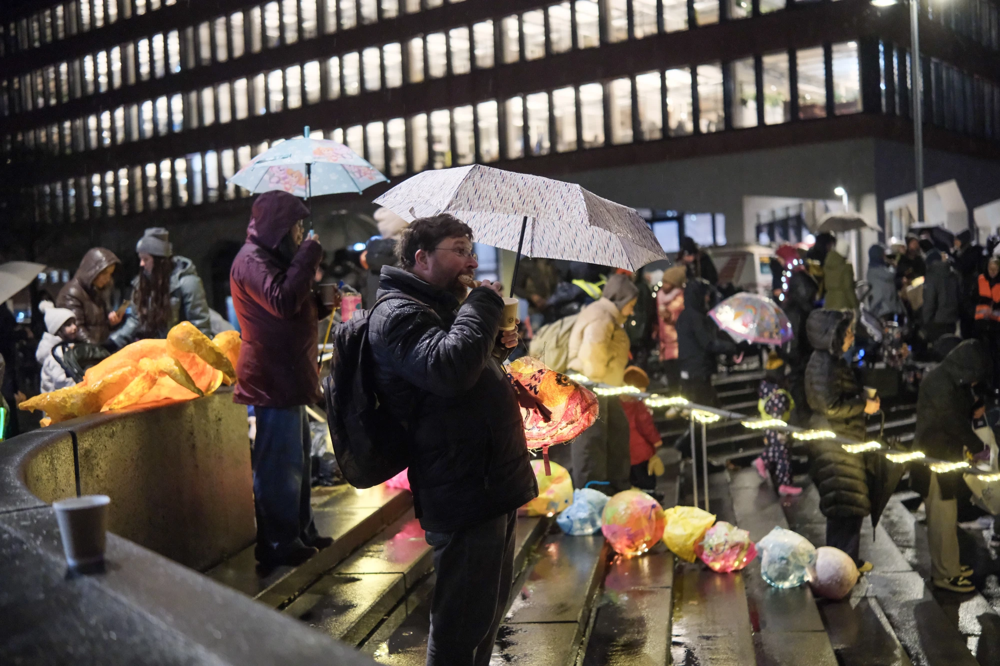 Menschen mit Regenschirmen stehen und sitzen auf Treppenstufen vor einem beleuchteten Gebäude bei Nacht.