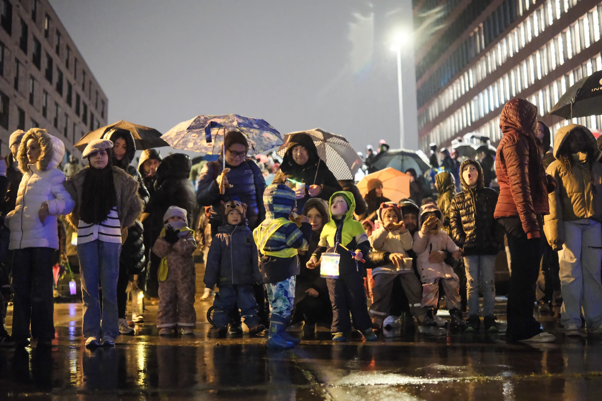 Gruppe von Kindern und Erwachsenen mit Regenschirmen und Laternen bei Nacht auf nassem Boden vor beleuchtetem Gebäude.
