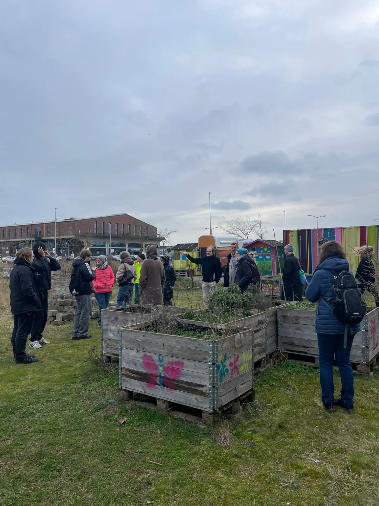 Mehrere Personen stehen auf einer Wiese zwischen großen, rechteckigen Pflanzkisten aus Holz. Einige Kisten sind mit bunten Motiven bemalt. Im Hintergrund ist ein langes Backsteingebäude zu sehen, daneben bunte vertikale Streifen auf einer Wand. Der Himmel ist bewölkt.