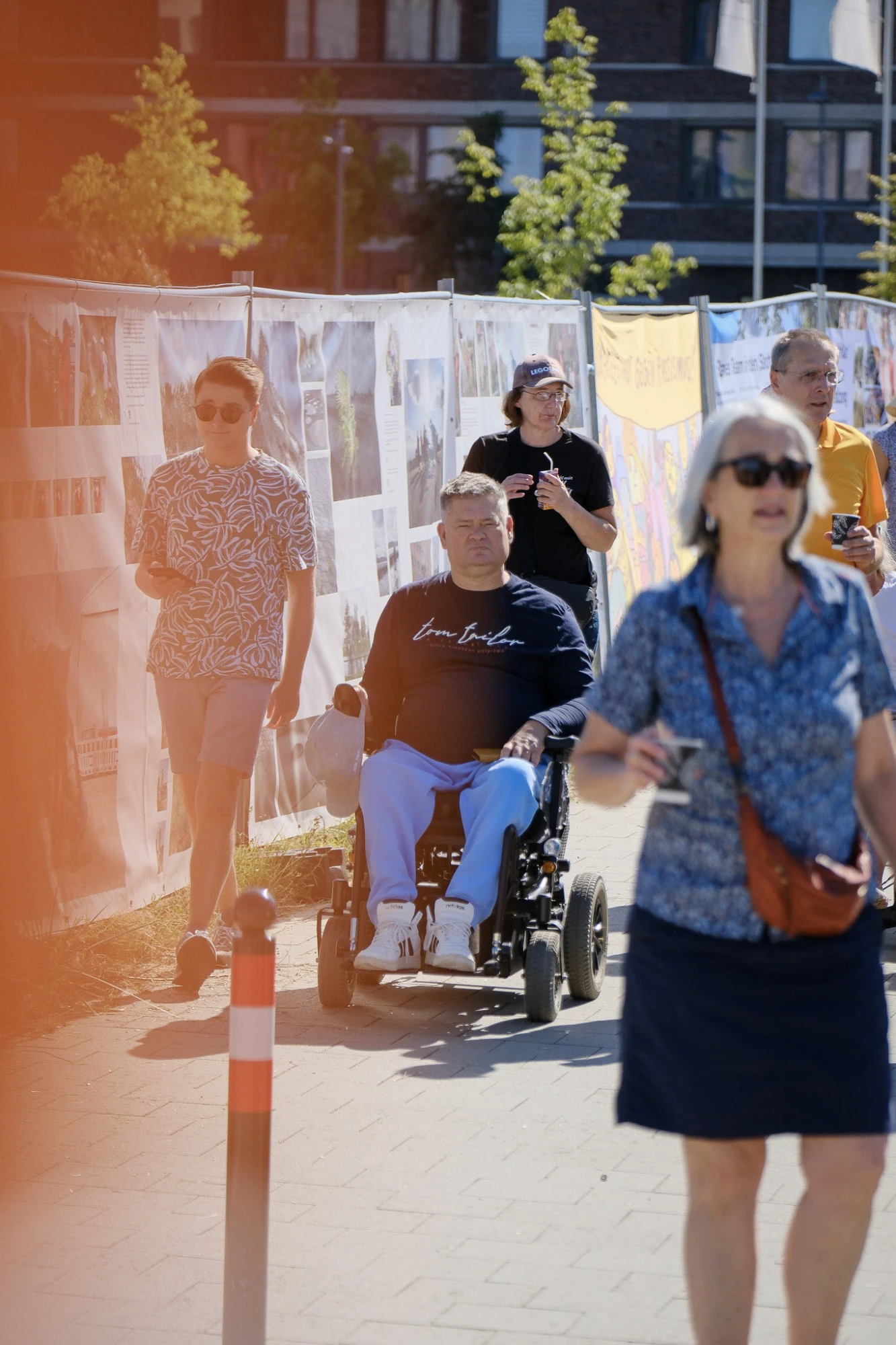 Im Vordergrund läuft eine Frau mit grauem Haar, Sonnenbrille und dunkelblauem Rock, die einen Becher in der Hand hält. Dahinter fährt ein Mann im Rollstuhl, der ein dunkelblaues Oberteil und hellblaue Hose trägt. Neben ihm gehen zwei Personen, eine mit gemustertem T-Shirt und kurzer Hose, die andere mit dunklem T-Shirt und Brille. Im Hintergrund sind weitere Menschen und eine lange Reihe von Plakaten oder Bannern zu sehen, die an einem Zaun befestigt sind. Die Szene spielt im Freien bei sonnigem Wetter.