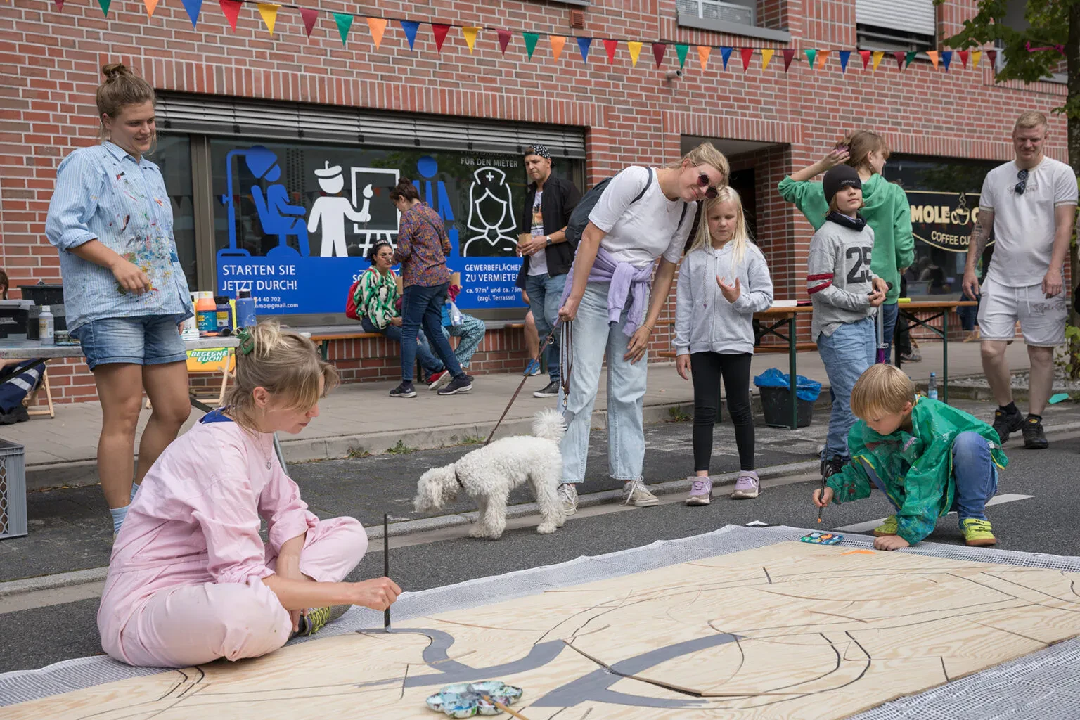 Auf der Straße findet ein Kinderkunstprojekt statt. Eine Frau malt auf einem Holzbrett, das auf der Straße liegt. Ein weiteres Kind malt auch mit. Andere Menschen, darunter auch ein Hund stehen drum herum und schauen zu.