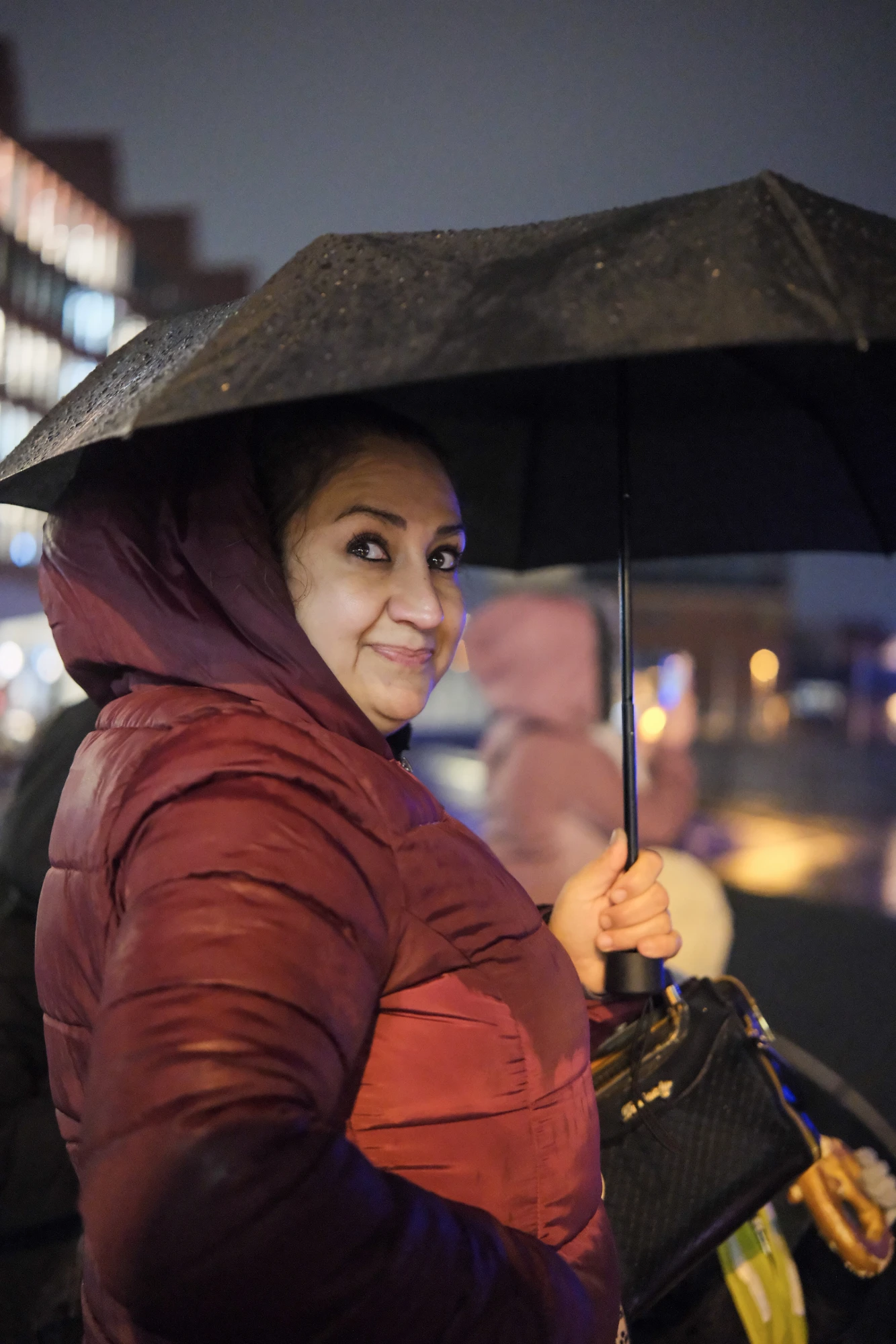 Person in roter Steppjacke hält bei Nacht einen geöffneten schwarzen Regenschirm, im Hintergrund unscharfe Lichter und weitere Personen.