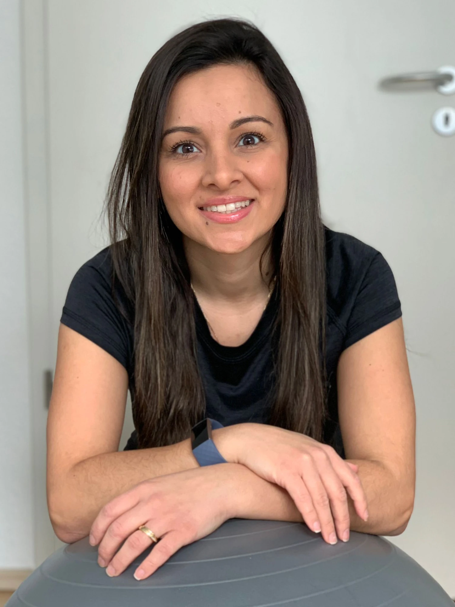 Eine Frau mit langen, glatten braunen Haaren tr&auml;gt ein schwarzes T-Shirt und sitzt mit verschr&auml;nkten Armen auf einem grauen Gymnastikball. An ihrem linken Handgelenk befindet sich ein Fitnessarmband, am rechten Ringfinger ein goldener Ring. Im Hintergrund ist eine helle T&uuml;r mit einem silberfarbenen T&uuml;rgriff und einem runden Schild zu sehen.