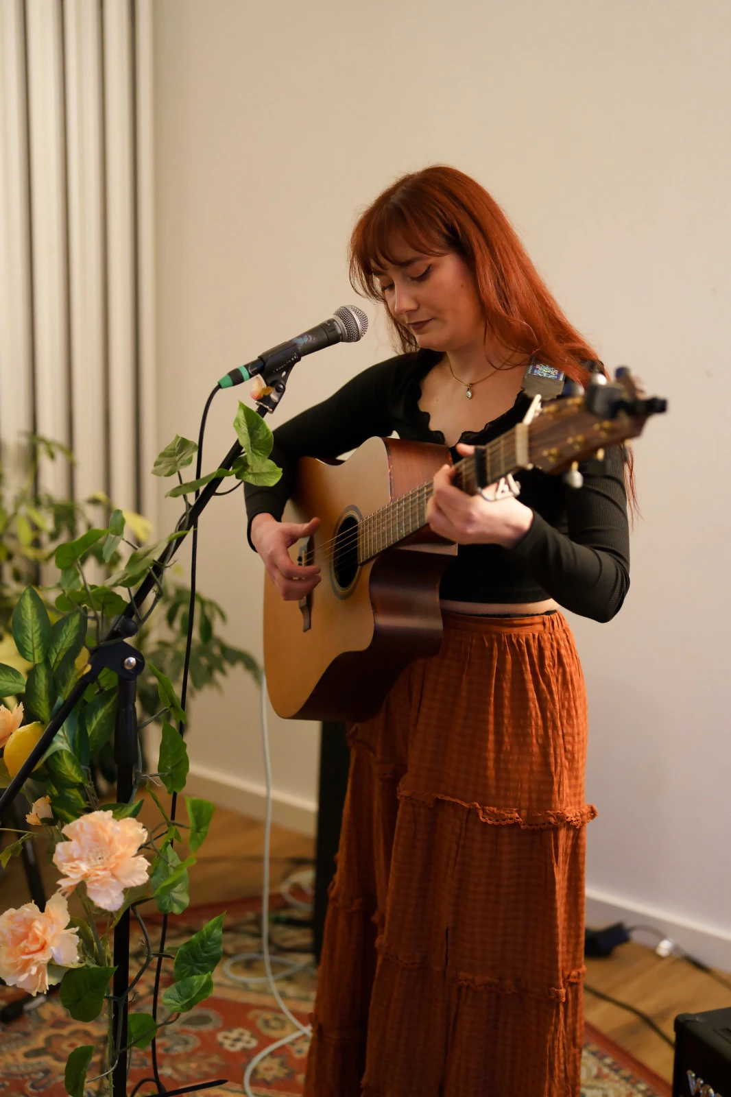 Die Musikerin Ragna Lemontree, mit langem rotem Haar spielt akustische Gitarre vor Mikrofon in einem Raum mit Pflanzen und Teppich