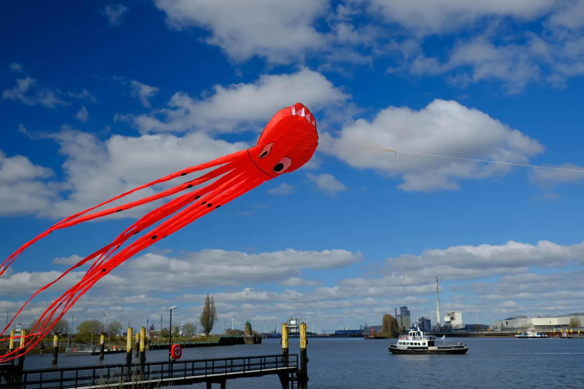 Zu sehen ist ein großer roter Drachen. Er sieht aus wie eine Kranke. Im Hintergrund sieht man die Weser und eine Fähre. Der Himmel ist blau.
