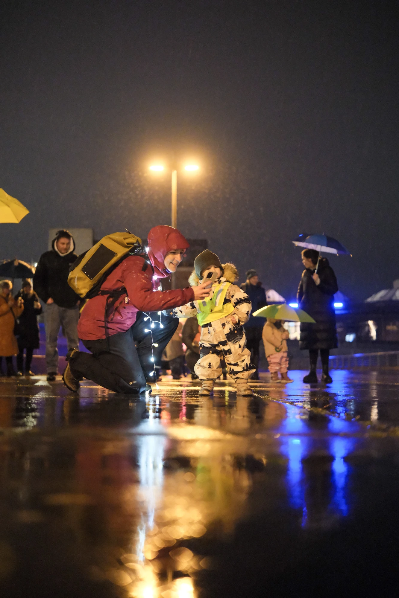 Zwei Kinder in bunter Regenkleidung knien auf nassem Boden, umgeben von mehreren Personen mit Regenschirmen bei Nacht.
