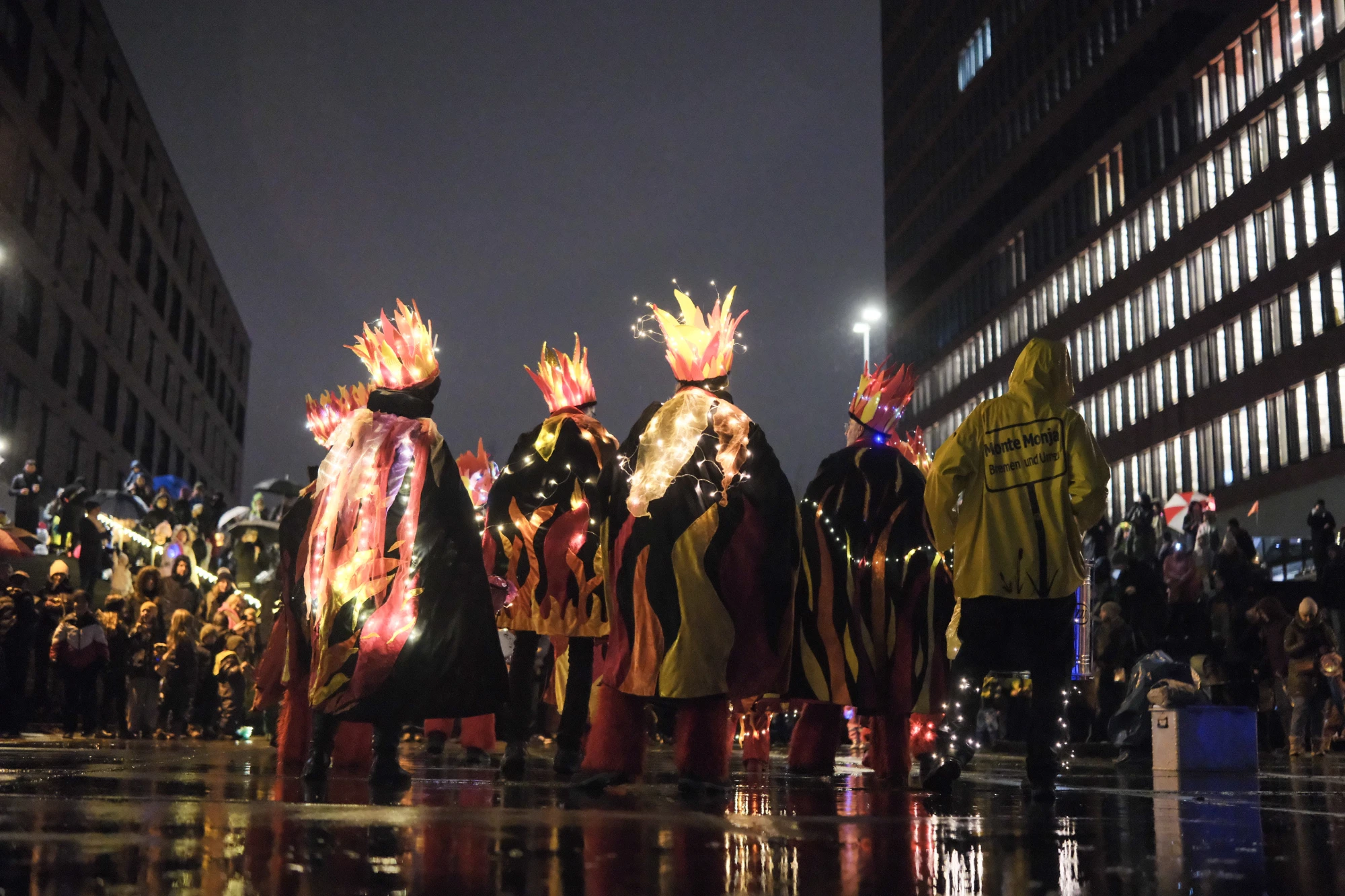 Gruppe von sechs Personen in leuchtenden Kostümen mit Flammenmotiven, eine Person spielt Trommel, nächtliche Stadtkulisse im Hintergrund.