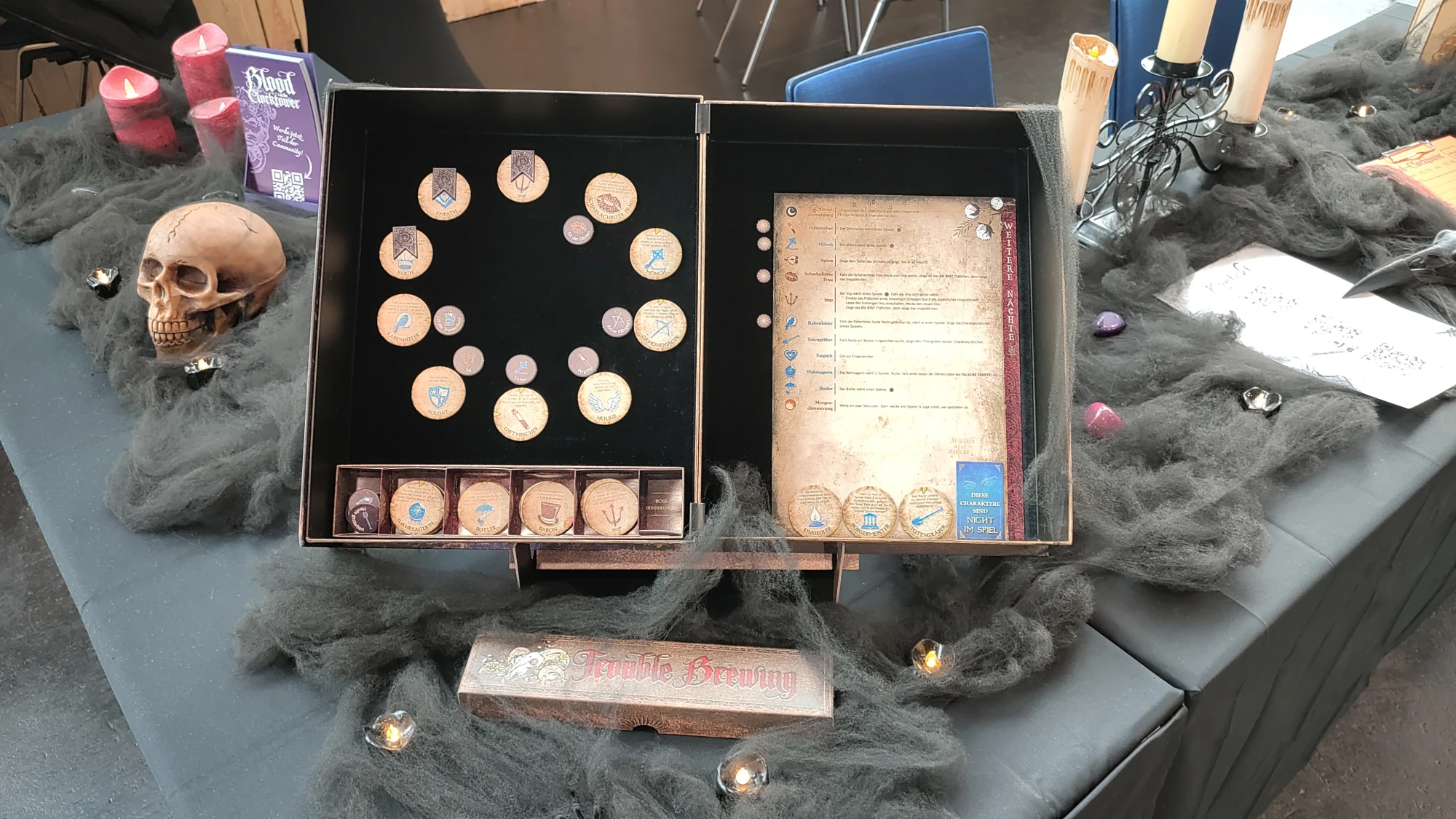Ein dekorativer Spieletisch mit einem ge&ouml;ffneten Spielkasten des Gesellschaftsspiels Blood on the Clocktower. Der Kasten zeigt verschiedene Holzmarker mit Symbolen und Beschriftungen, sowie eine &Uuml;bersichtstafel mit Spielregeln. Um den Kasten herum liegen schwarze Deko-Spinnweben, kleine LED-Teelichter, ein k&uuml;nstlicher Sch&auml;del, rote Kerzen und ein Kerzenst&auml;nder mit wei&szlig;en Kerzen, die eine mystische, Halloween-&auml;hnliche Atmosph&auml;re erzeugen.