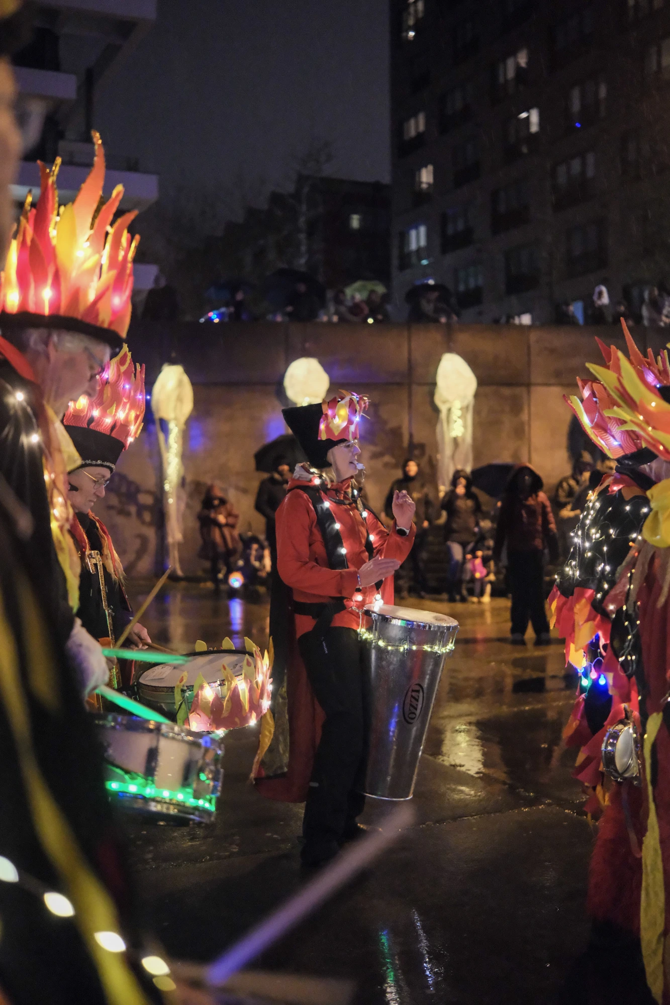 Person in rotem Kostüm mit leuchtender Kopfbedeckung spielt Trommel bei nächtlicher Veranstaltung im Freien.