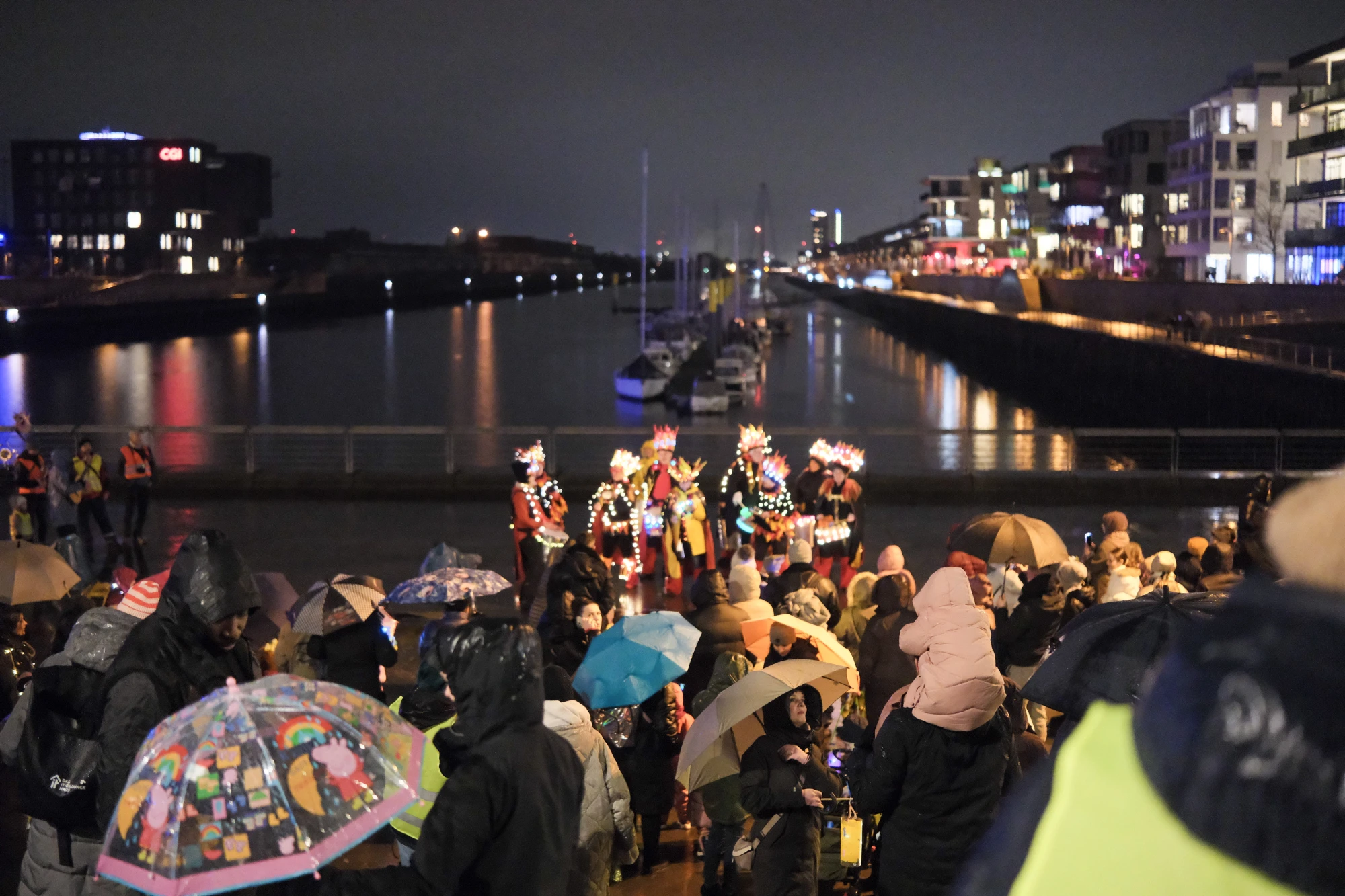 Menschenmenge mit Regenschirmen und Kostümen an einem Hafen bei Nacht, im Hintergrund beleuchtete Gebäude und Boote im Wasser.