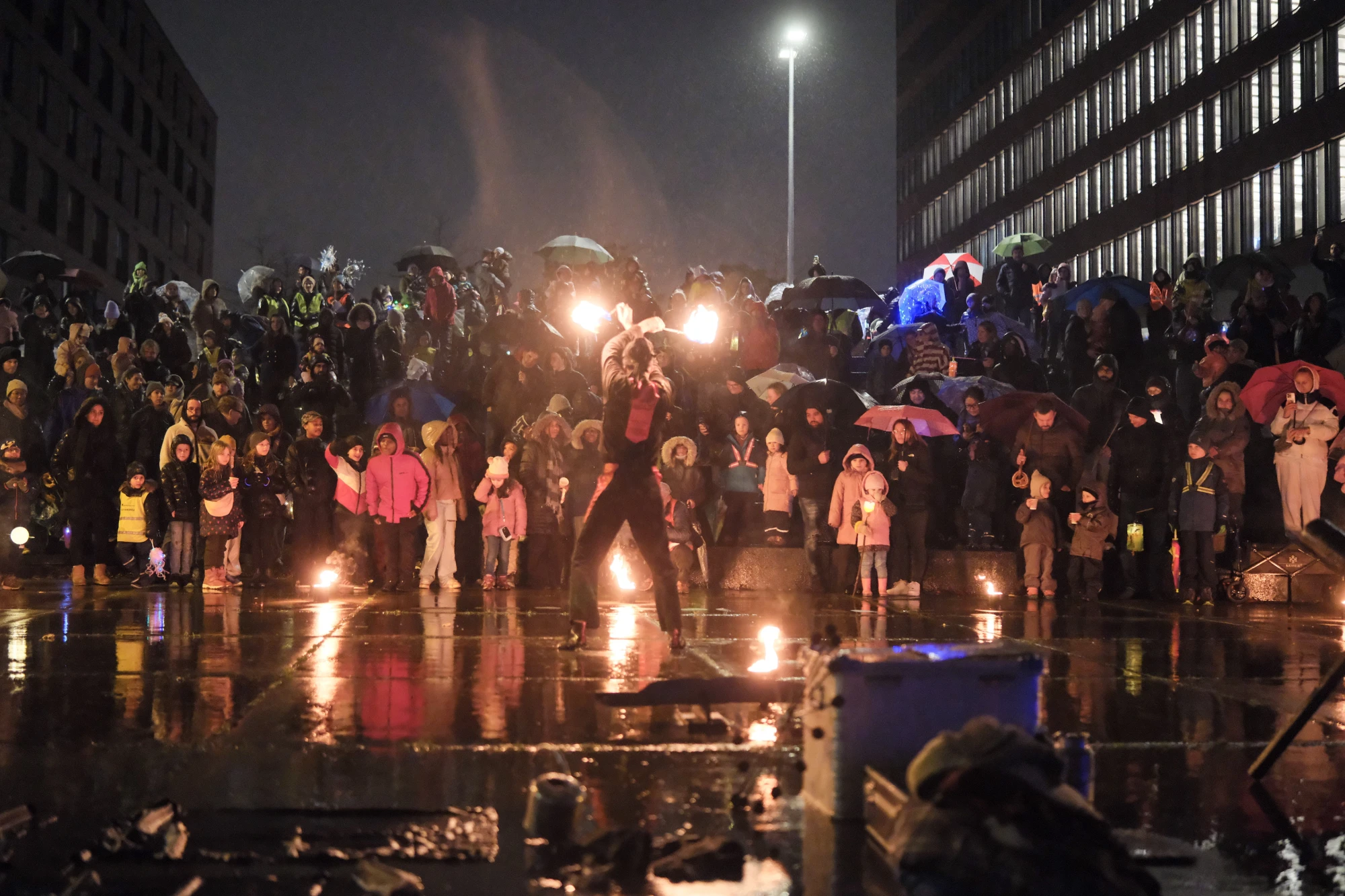 Feuerkünstler in Bewegung vor einer großen Menschenmenge bei Nacht, die mit Regenschirmen und warmen Kleidern zuschaut.
