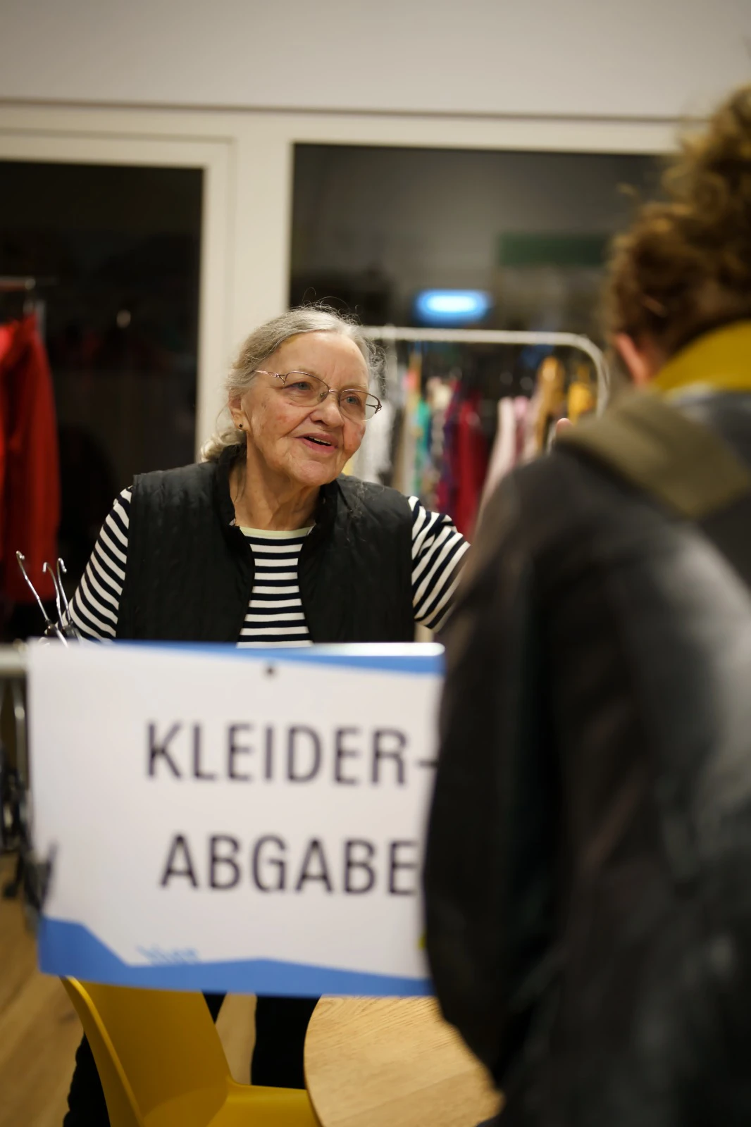 Person mit gestreiftem Oberteil steht hinter einem Tisch mit Schild 'KLEIDER ABGABE' und spricht mit einer anderen Person mit Rucksack