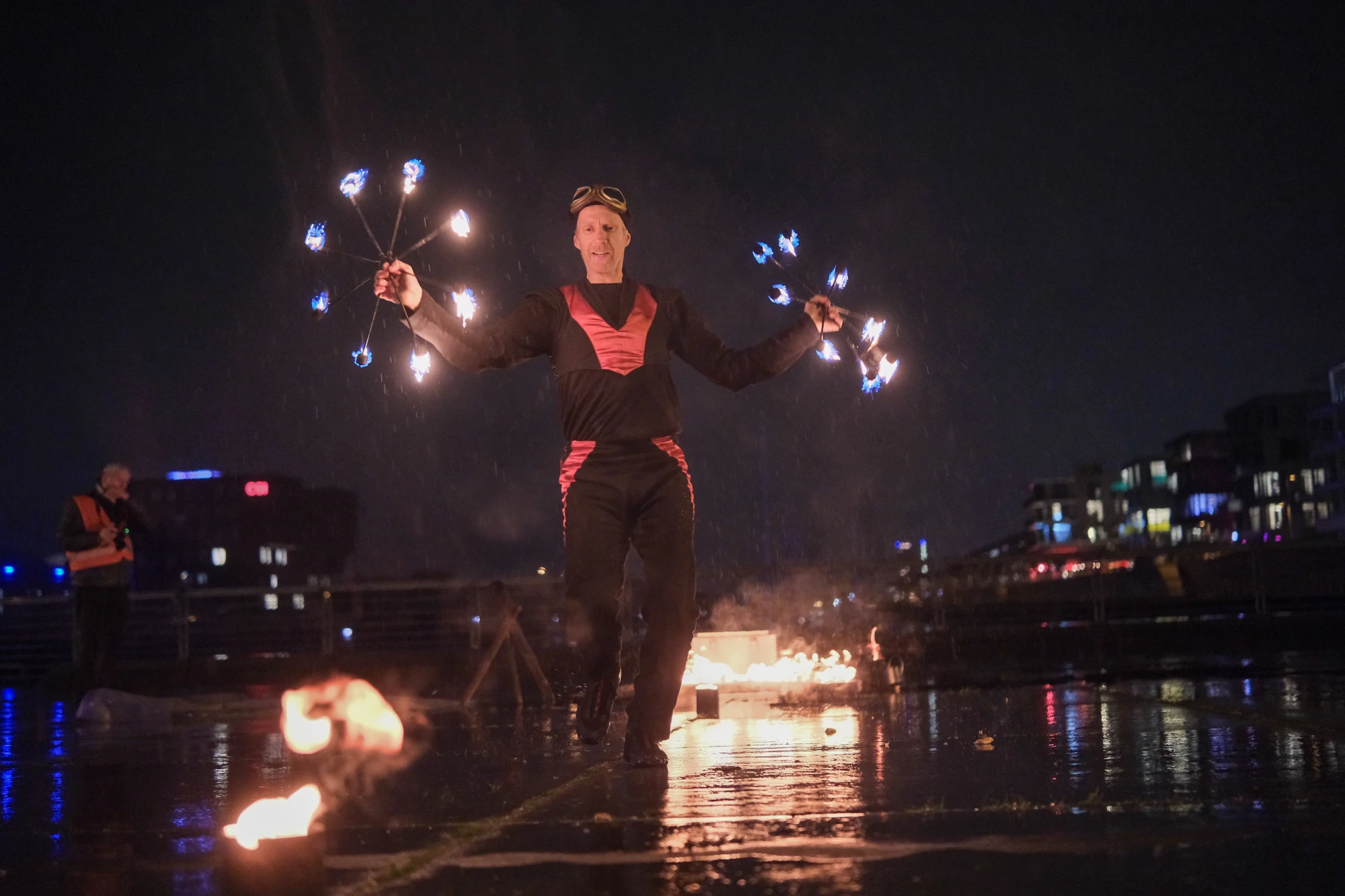 Person in dunkler Kleidung mit reflektierenden Streifen hält brennende Laterne über nassem Boden bei Nacht.