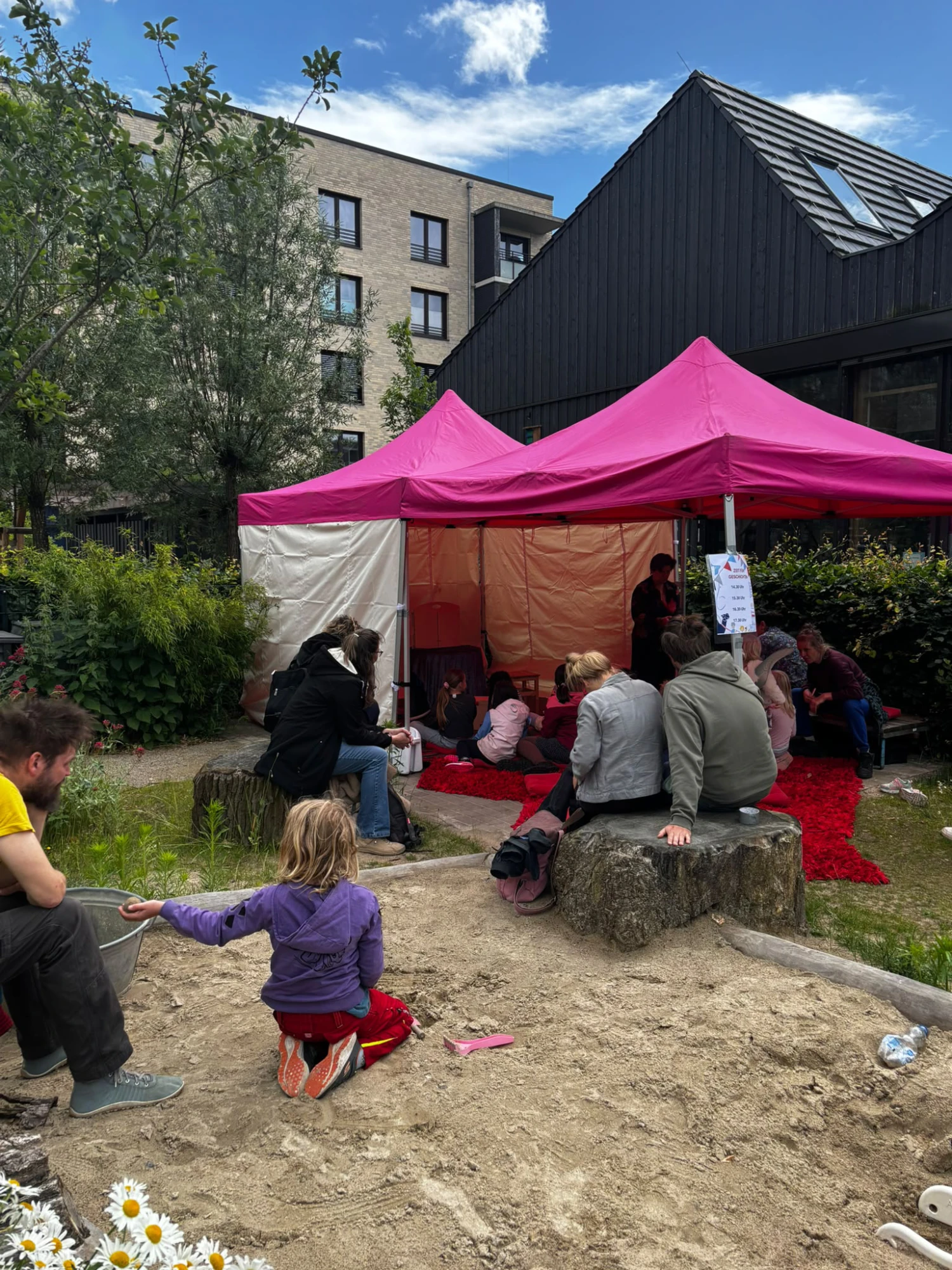 Kinder und Erwachsene sitzen und spielen unter einem pinkfarbenen Pavillon in einem Garten mit Sandkasten vor einem modernen Wohngebäude