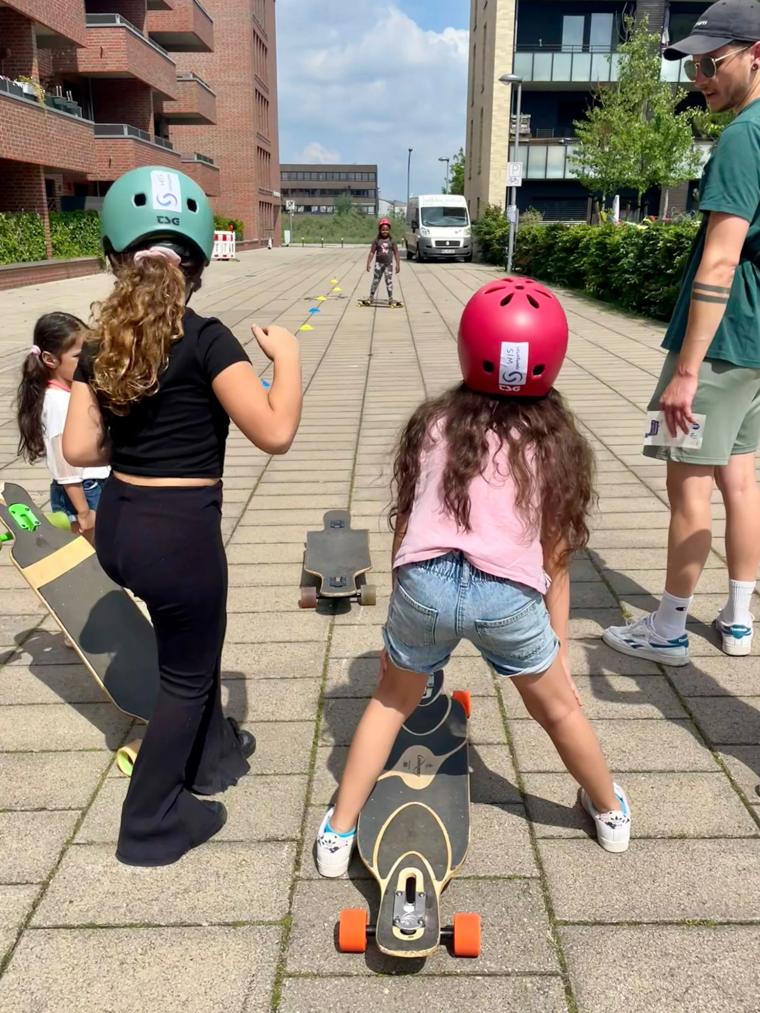 Mehrere Kinder mit Helmen stehen auf Longboards auf einem gepflasterten Weg, ein Erwachsener steht daneben