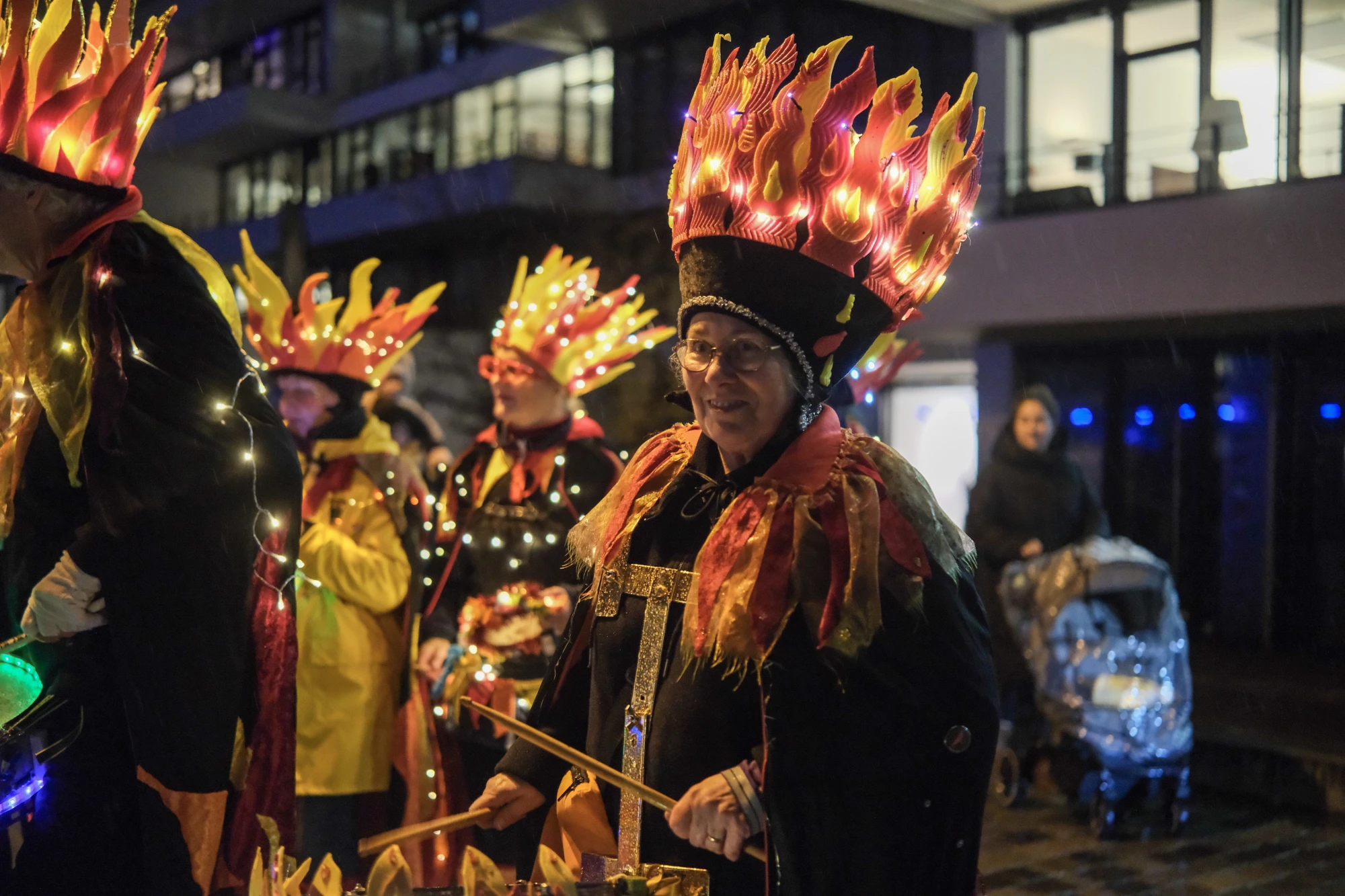 Gruppe von Menschen in schwarzen Kostümen mit leuchtenden, flammenförmigen Kopfbedeckungen, einige spielen Trommeln mit grünen und blauen Lichtern.