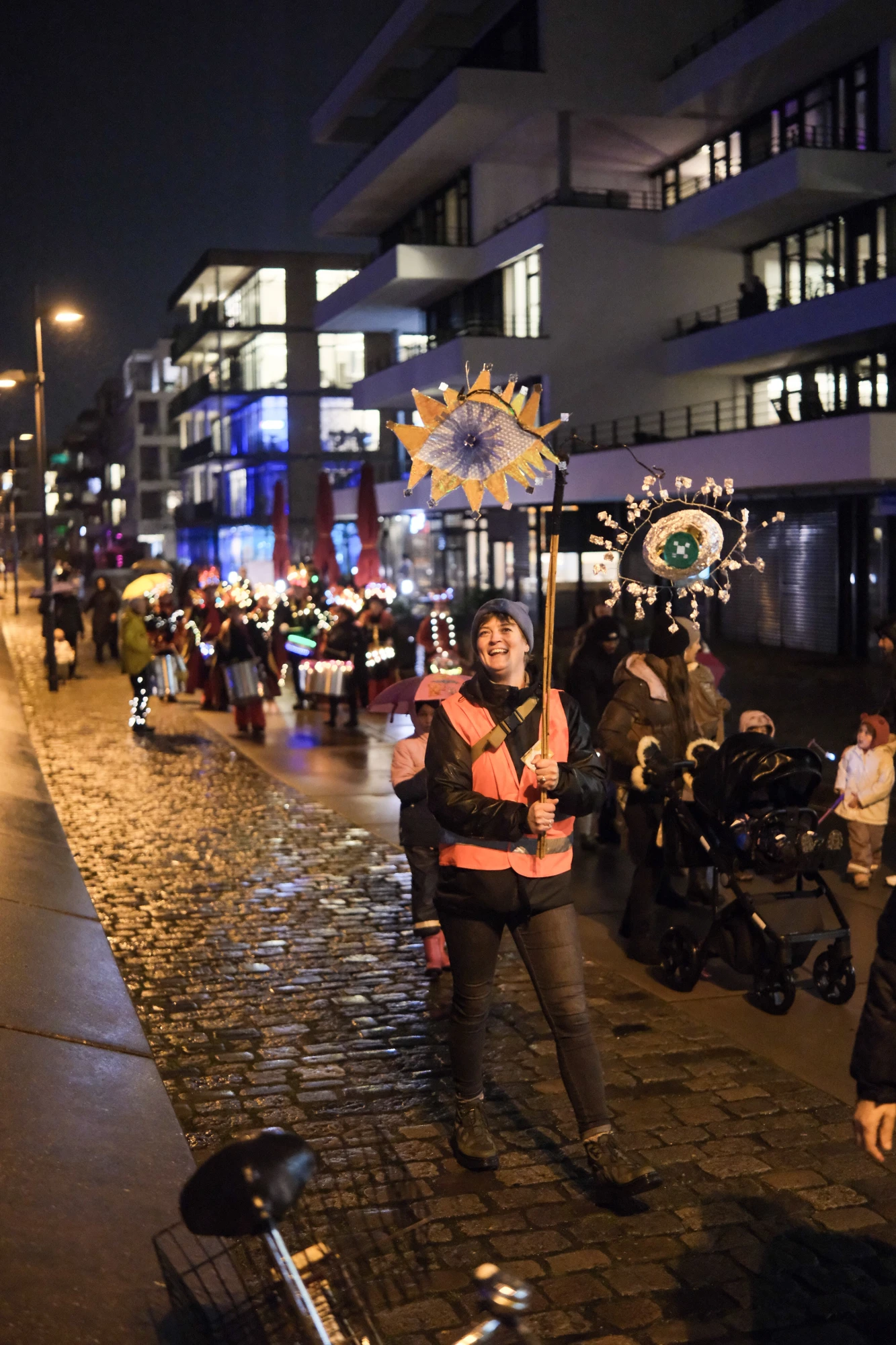 Person mit orangefarbener Warnweste trägt eine Laterne mit blau-gelbem Muster bei einem nächtlichen Umzug auf Kopfsteinpflasterstraße.
