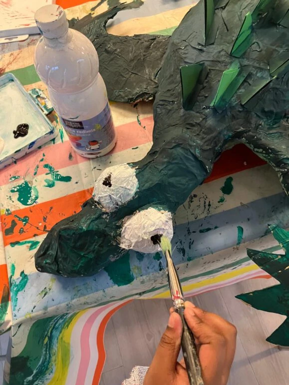 Ein gebasteltet Krokodil liegt auf einem bunten Tisch. Eine Hand mit Pinsel bemalt die Augen des Krokodils.