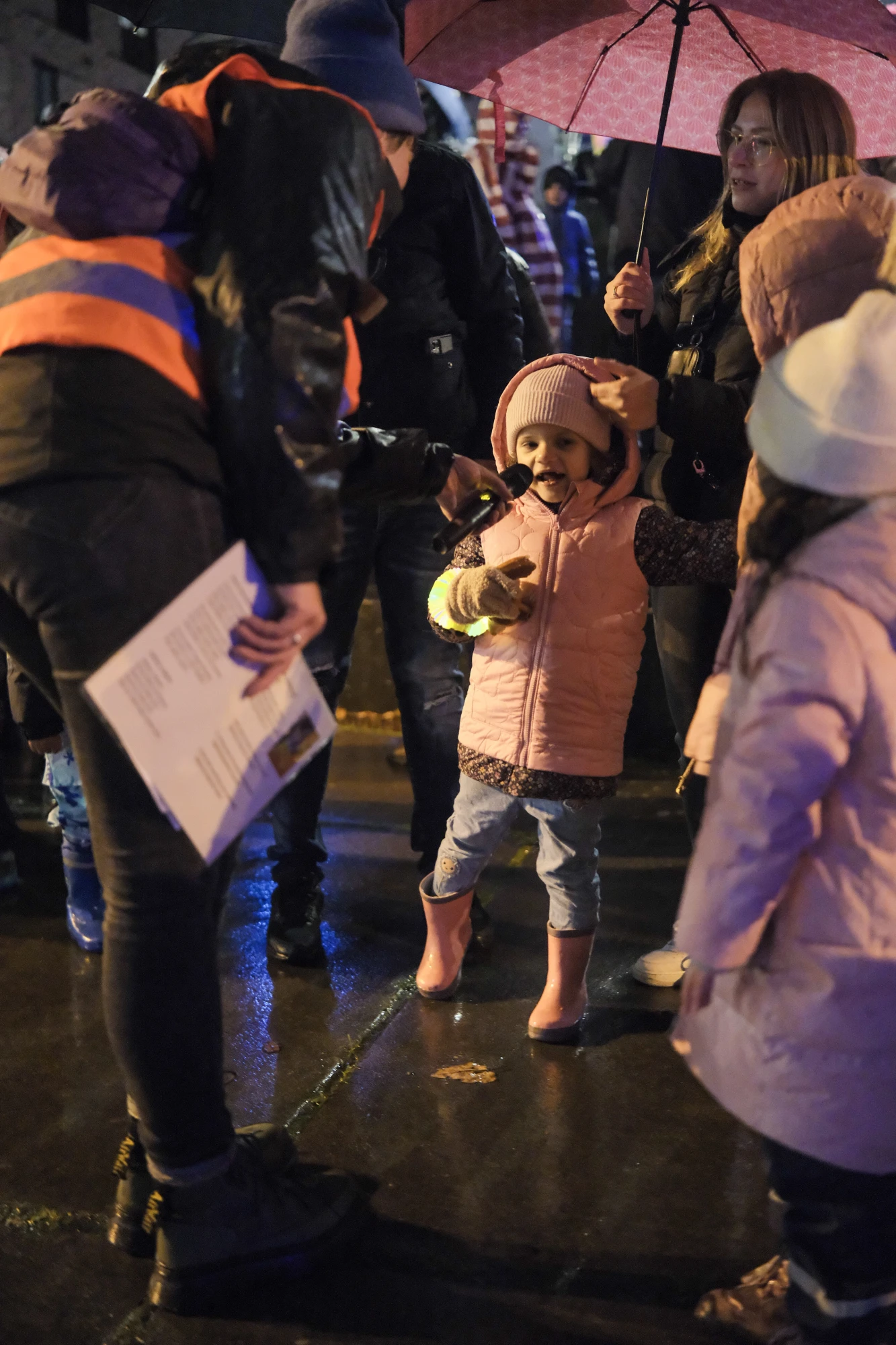 Kind in rosa Jacke und Gummistiefeln steht auf nassem Boden, umgeben von mehreren Personen, eine hält einen Regenschirm.
