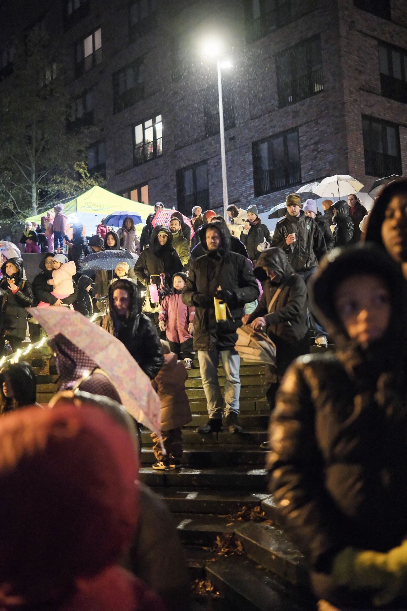 Menschenmenge auf einer Treppe bei Nacht, einige mit Regenschirmen, im Hintergrund ein beleuchteter Straßenlaterne und ein Zelt.