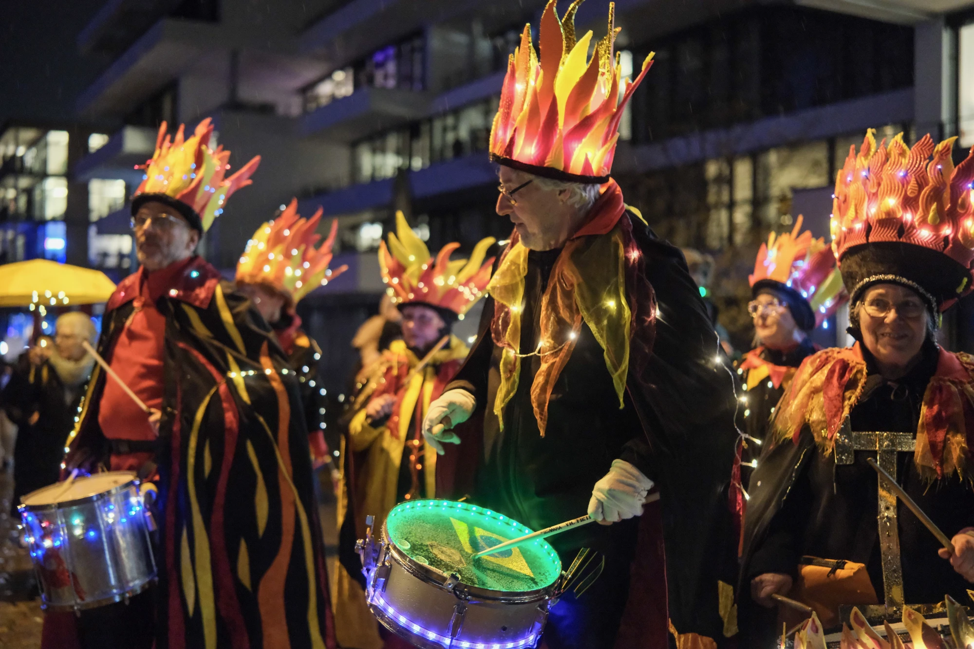Gruppe von Menschen in schwarzen Kostümen mit leuchtenden, flammenförmigen Kopfbedeckungen, einige spielen Trommeln mit grünen und blauen Lichtern.