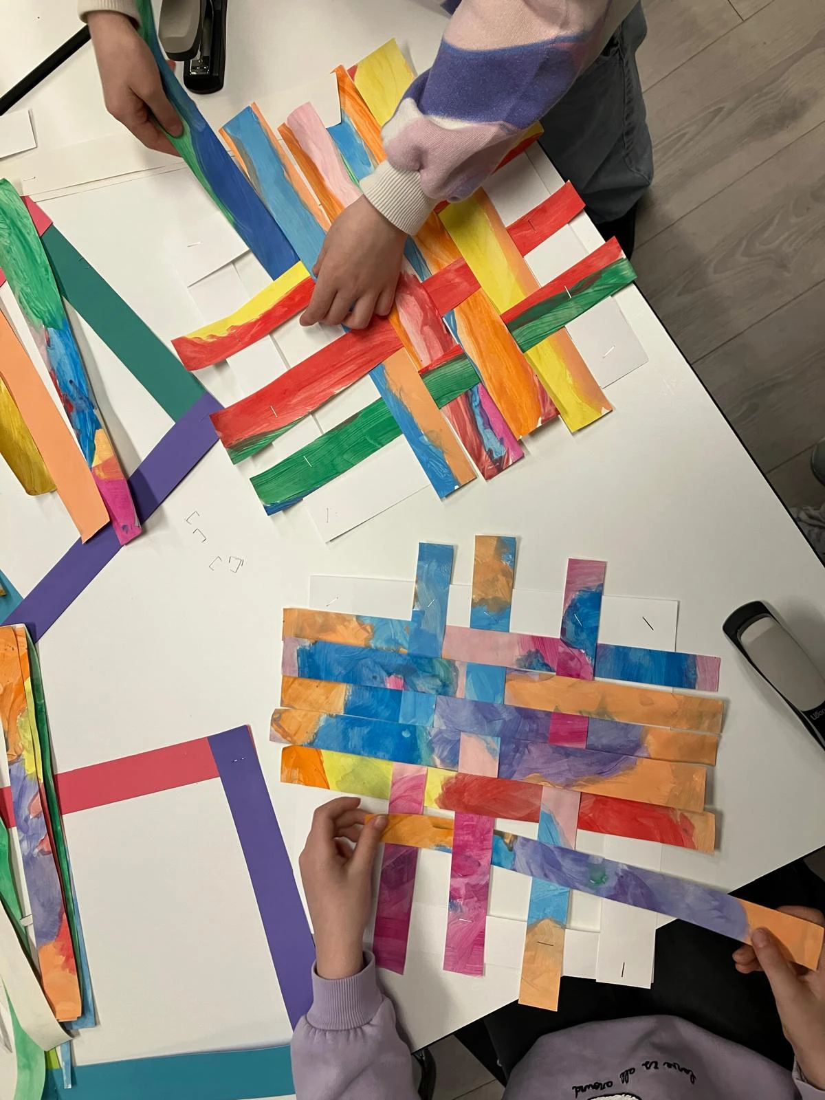 Auf einem Tisch liegen viele bunte Papierstreifen. Sie werden von Kinderhänden eingefädelt. Sie weben mit den Streifen.