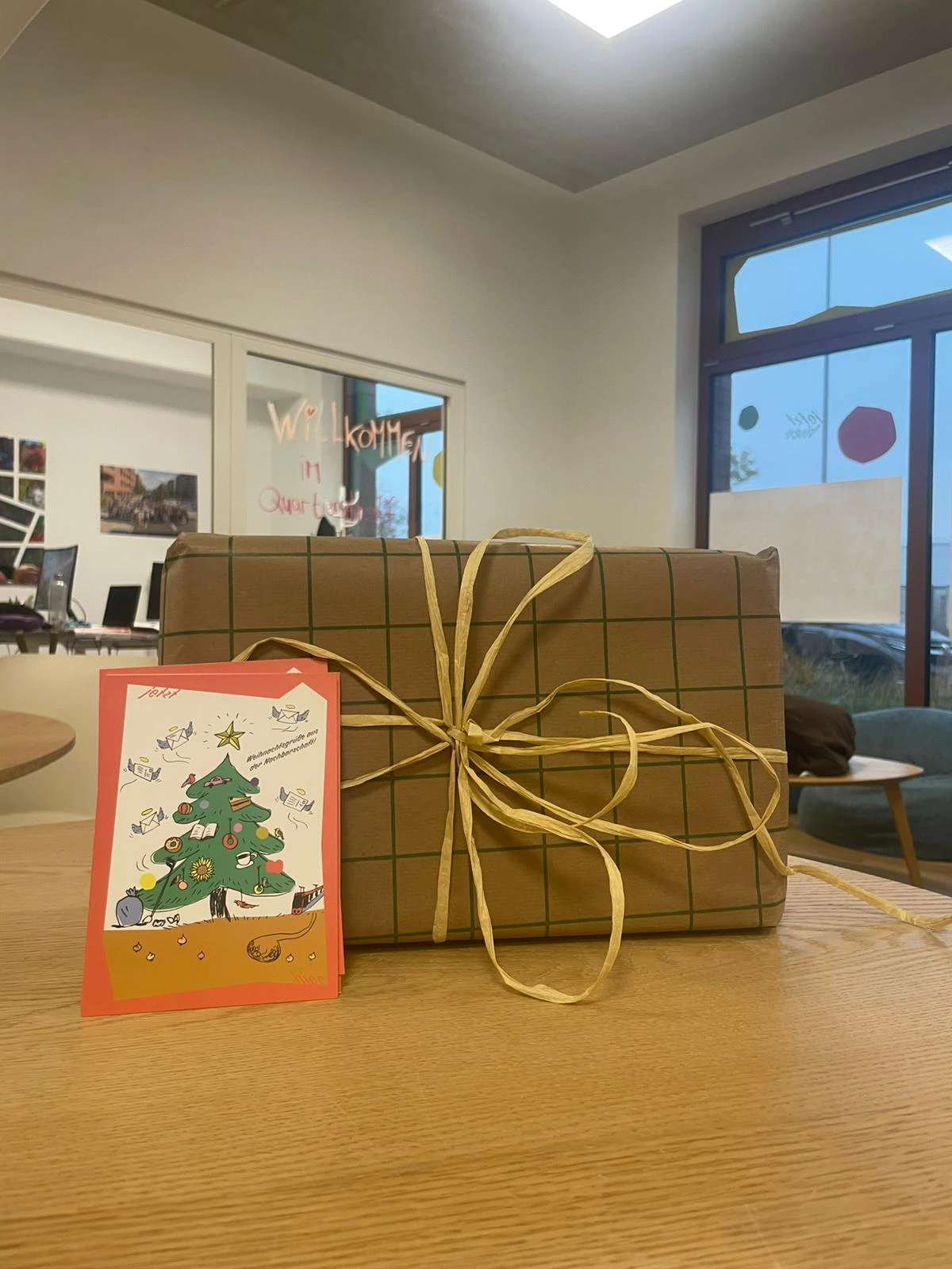 Zu sehen ist ein eingepacktes Geschenk mit einer Weihnachskarte auf einem Tisch. Im Hintergrund sind die R&auml;umlichkeiten des Quartierstreff zu sehen.
