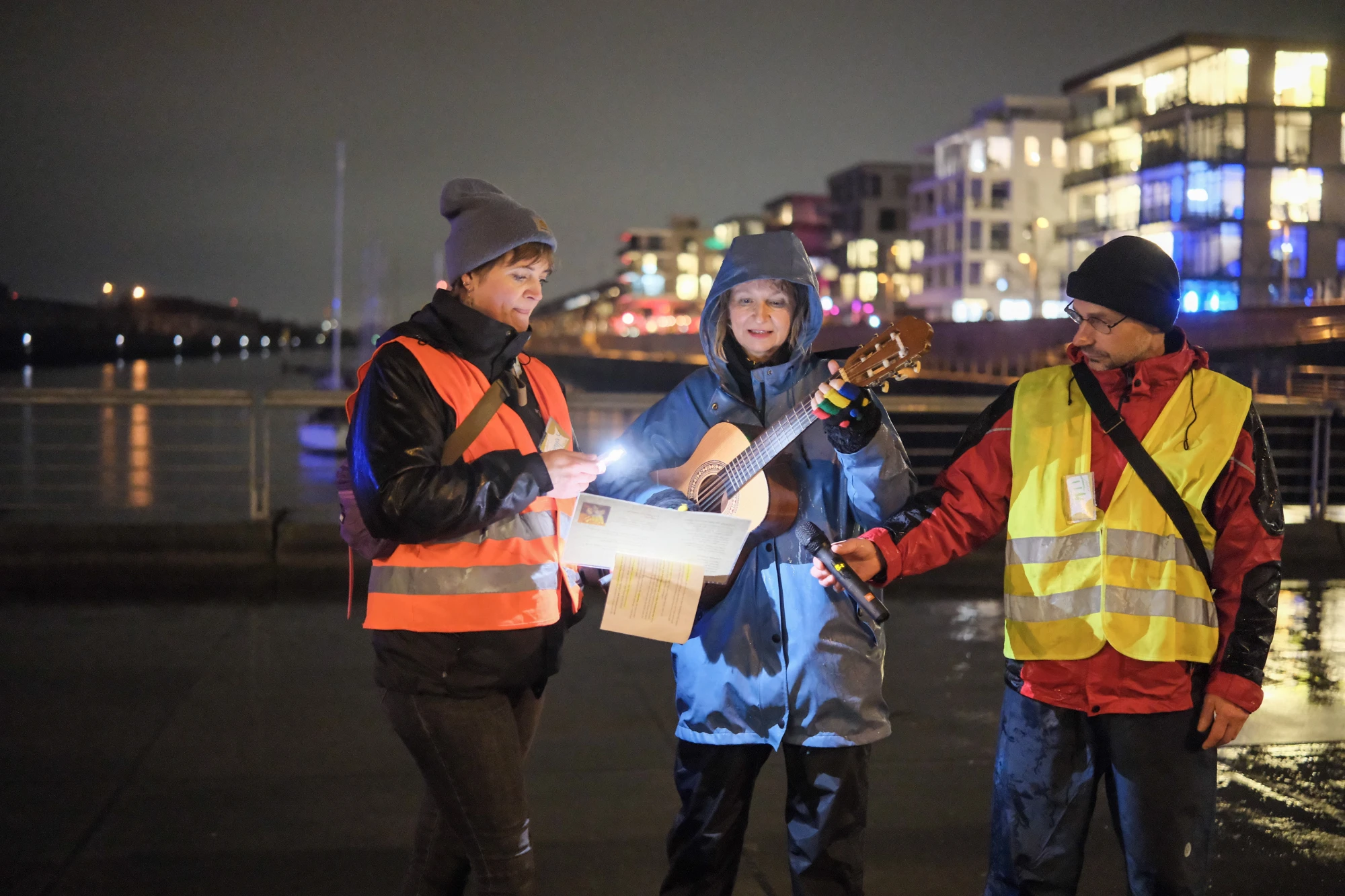 Drei Personen in Warnwesten stehen nachts an einem Wasserweg, eine spielt Gitarre, eine hält ein Blatt Papier, die dritte hält ein Mikrofon.