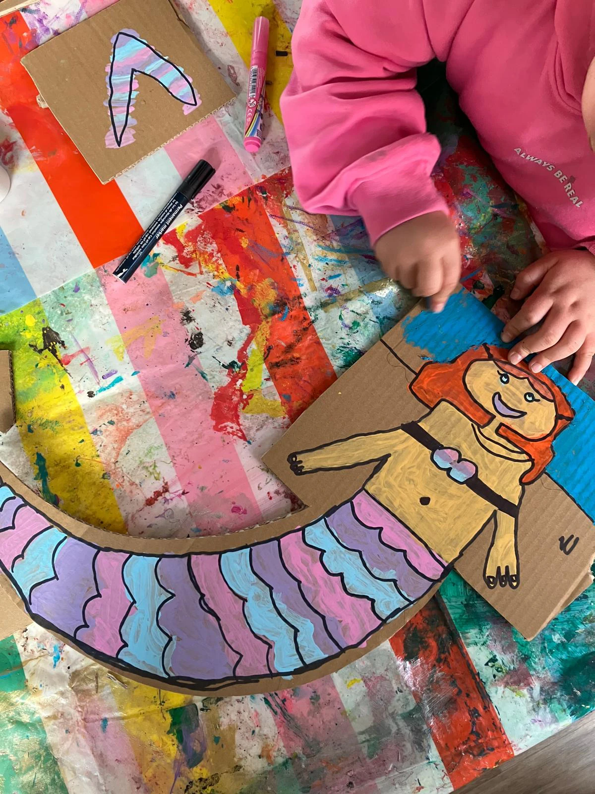 Eine bunt ausgemalte Meerjungfrau aus Pappe liegt auf einer bunten Tischdecke, die komplett mit Farbe voll ist. Ein Kind, dessen H&auml;nde zu sehen sind, malt gerade den HIntergrund blau an.