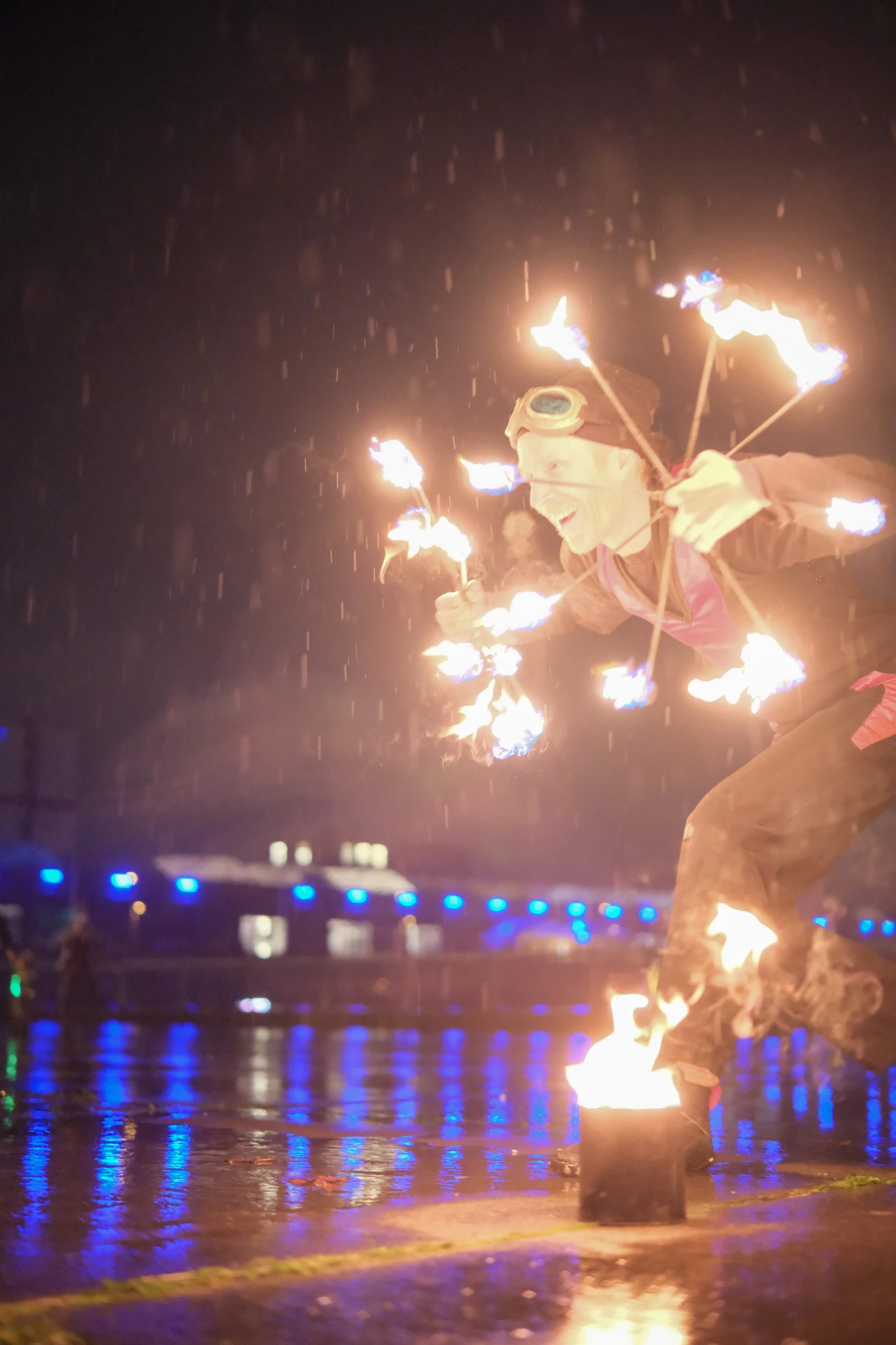 Person in schwarzem Outfit mit roten Akzenten jongliert nachts mit zwei brennenden Feuerreifen auf nassem Boden vor dunklem Hintergrund.