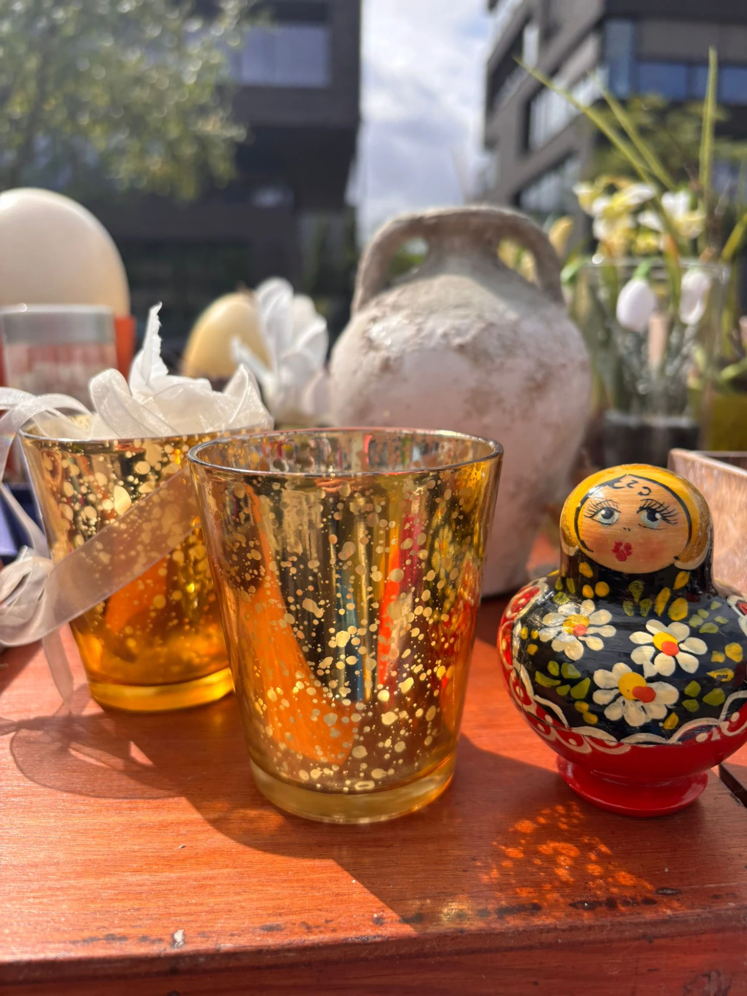 Zwei goldene Gl&auml;ser mit Sprenkeln, eine bemalte Matroschka-Puppe mit Blumenmuster und eine antike Tonvase auf einem Holztisch im Freien