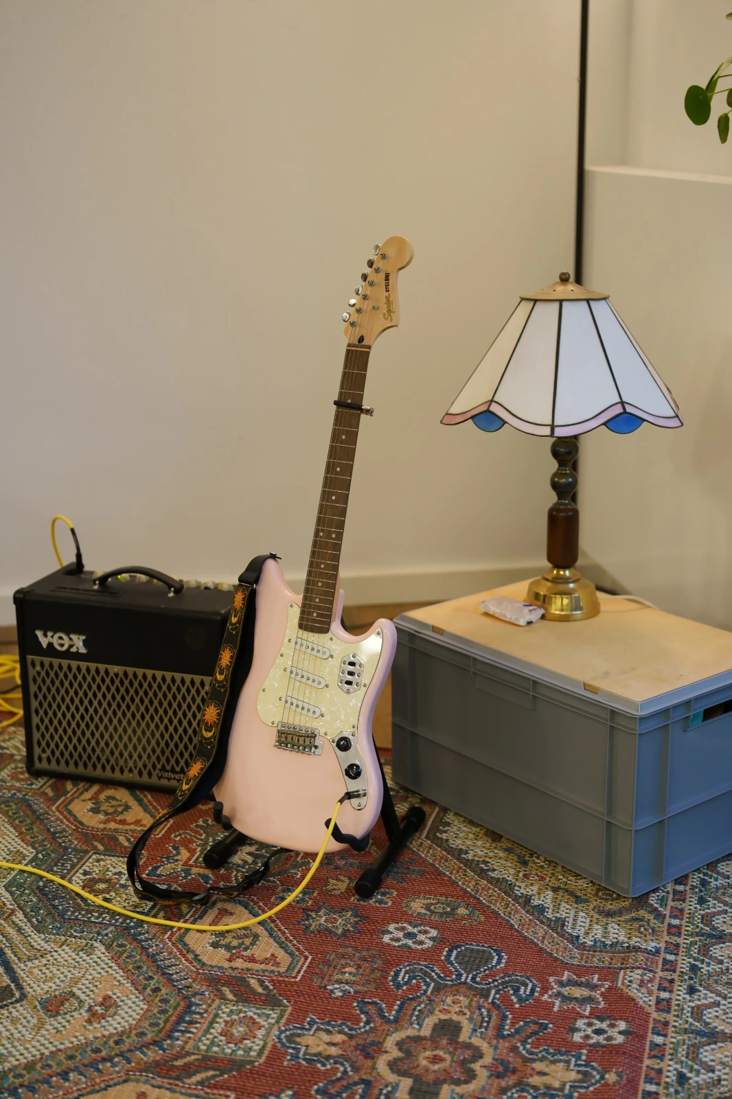 Hellrosa E-Gitarre lehnt an einem Vox-Verstärker auf einem gemusterten Teppich neben einem grauen Kasten mit einer Tischlampe