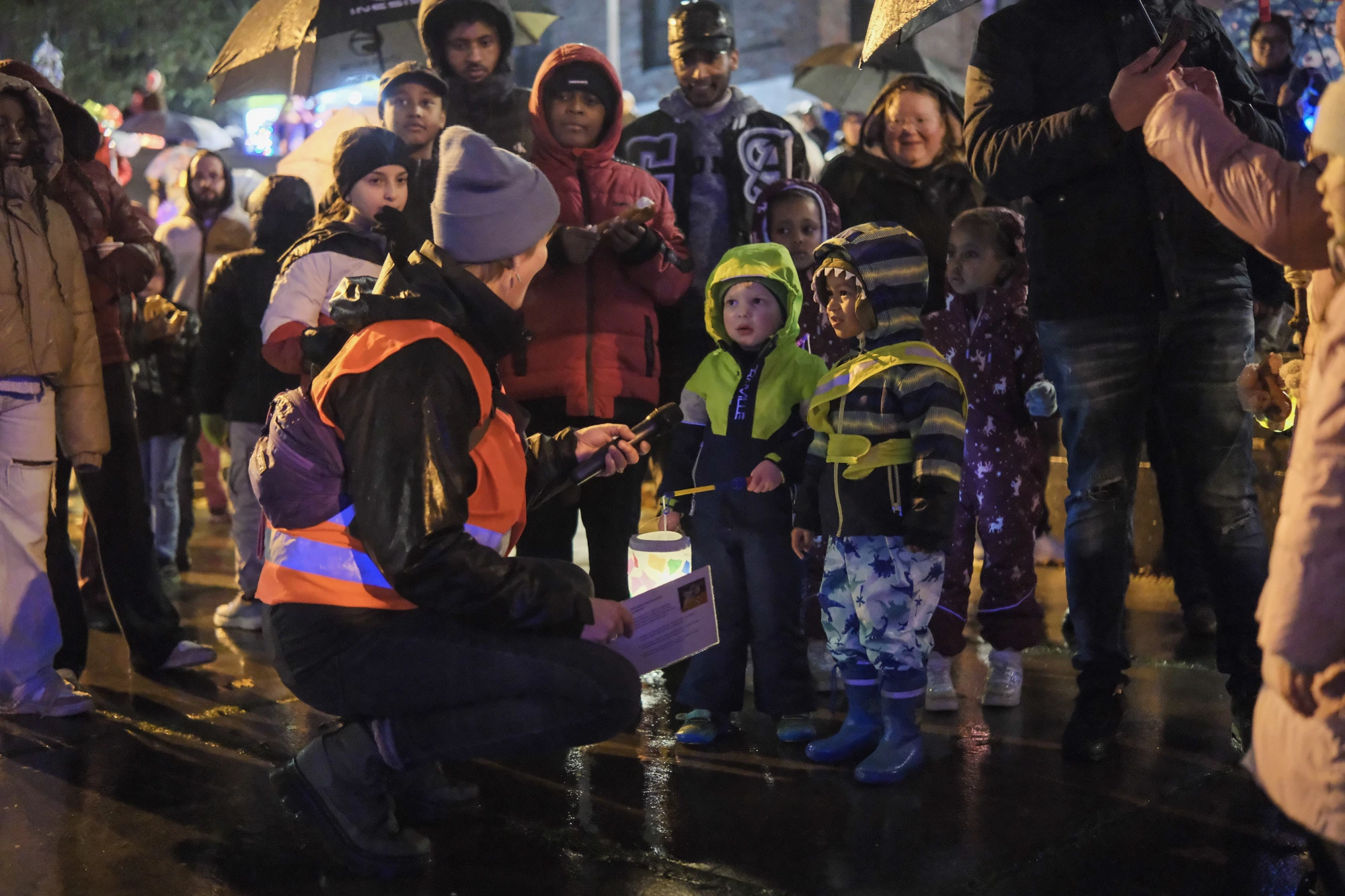 Person mit grauer Mütze und orangefarbener Warnweste kniet vor mehreren Kindern mit Laternen auf nassem Straßenbelag bei Nacht.