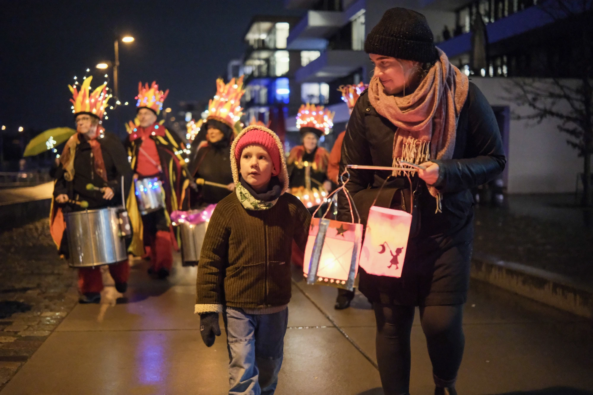 Eine Gruppe von Menschen mit leuchtenden Kronen und Laternen, vorne ein Kind und eine Erwachsene mit Laternen, nachts auf einem Gehweg.