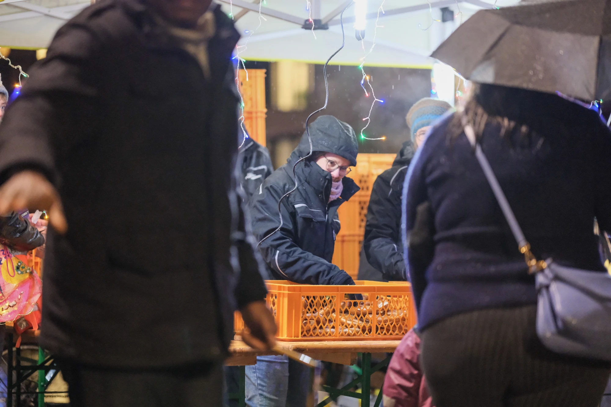 Personen stehen an einem Marktstand mit orangen Kunststoffkisten, eine Person hält einen Regenschirm, es ist Nacht und Lichterketten sind sichtbar.