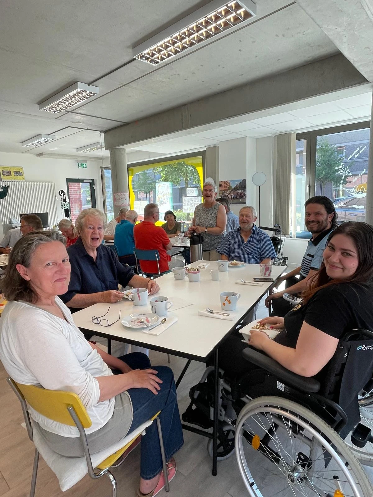 Auf dem Bild sind viele Leute zu sehen. Sie treffen sich zum Nachbarschaftscafé. Auf den Tischen stehen Teller und Tassen. Einige lachen freundlich in die Kamera.