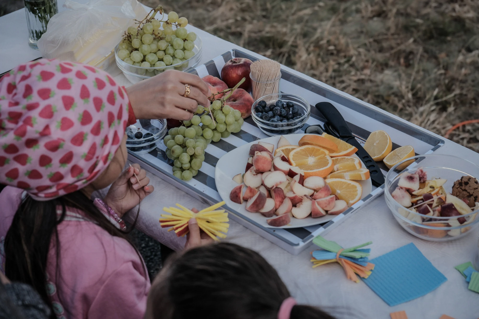 Kinder sitzen an einem Tisch mit Tellern voller Obst und Snacks, eine Hand greift nach einer Traube