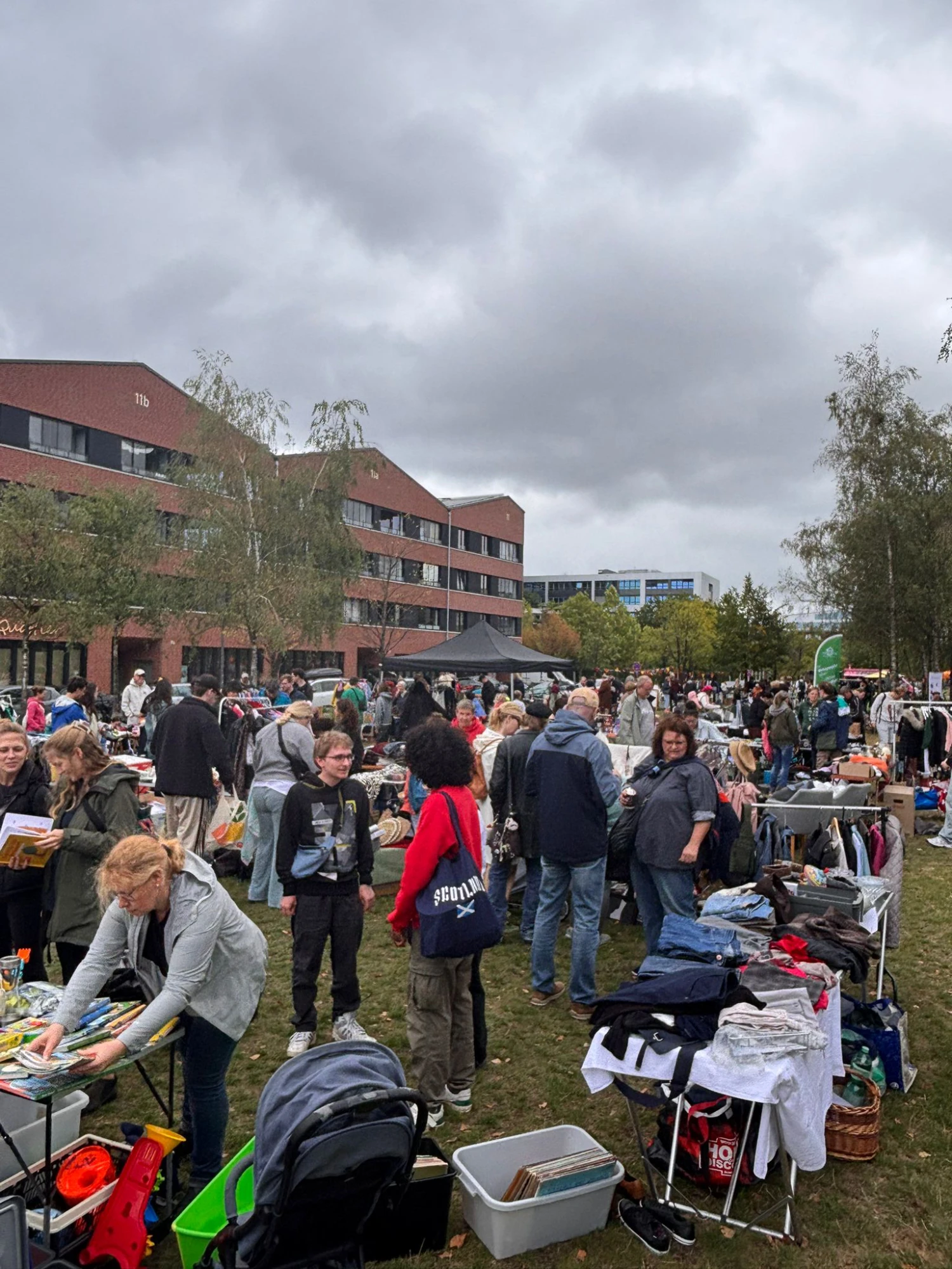 Menschen auf einem Flohmarkt im Freien vor mehrstöckigen Backsteingebäuden unter bewölktem Himmel