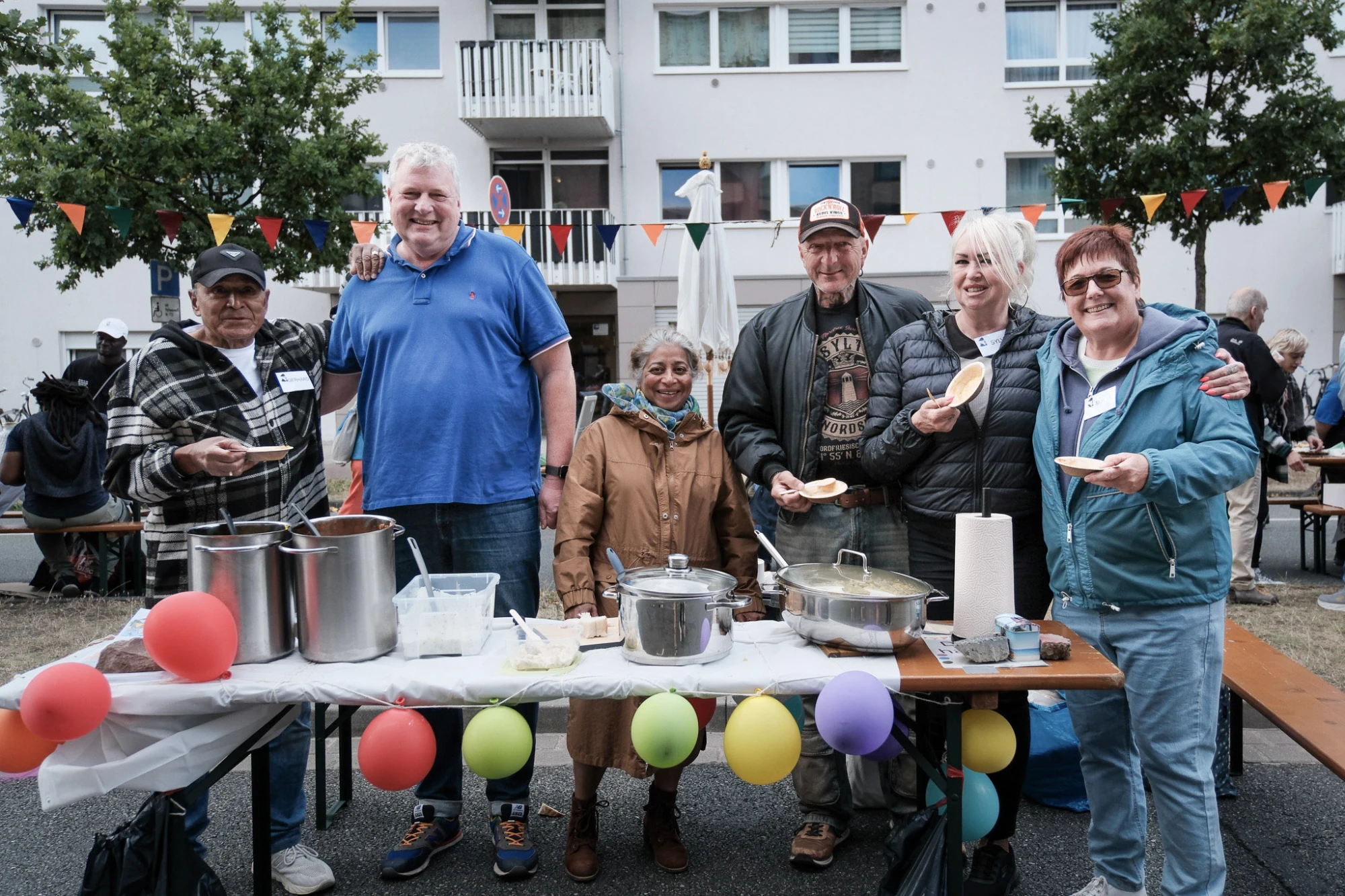 Sechs Personen stehen hinter einem mit bunten Luftballons geschmückten Tisch mit großen Töpfen und Servierutensilien vor einem Wohnhaus