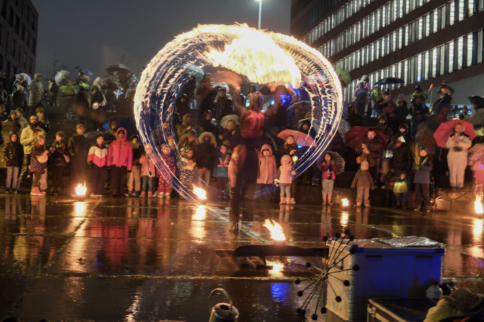 Feuerkünstler in Bewegung vor einer großen Menschenmenge bei Nacht, die mit Regenschirmen und warmen Kleidern zuschaut.
