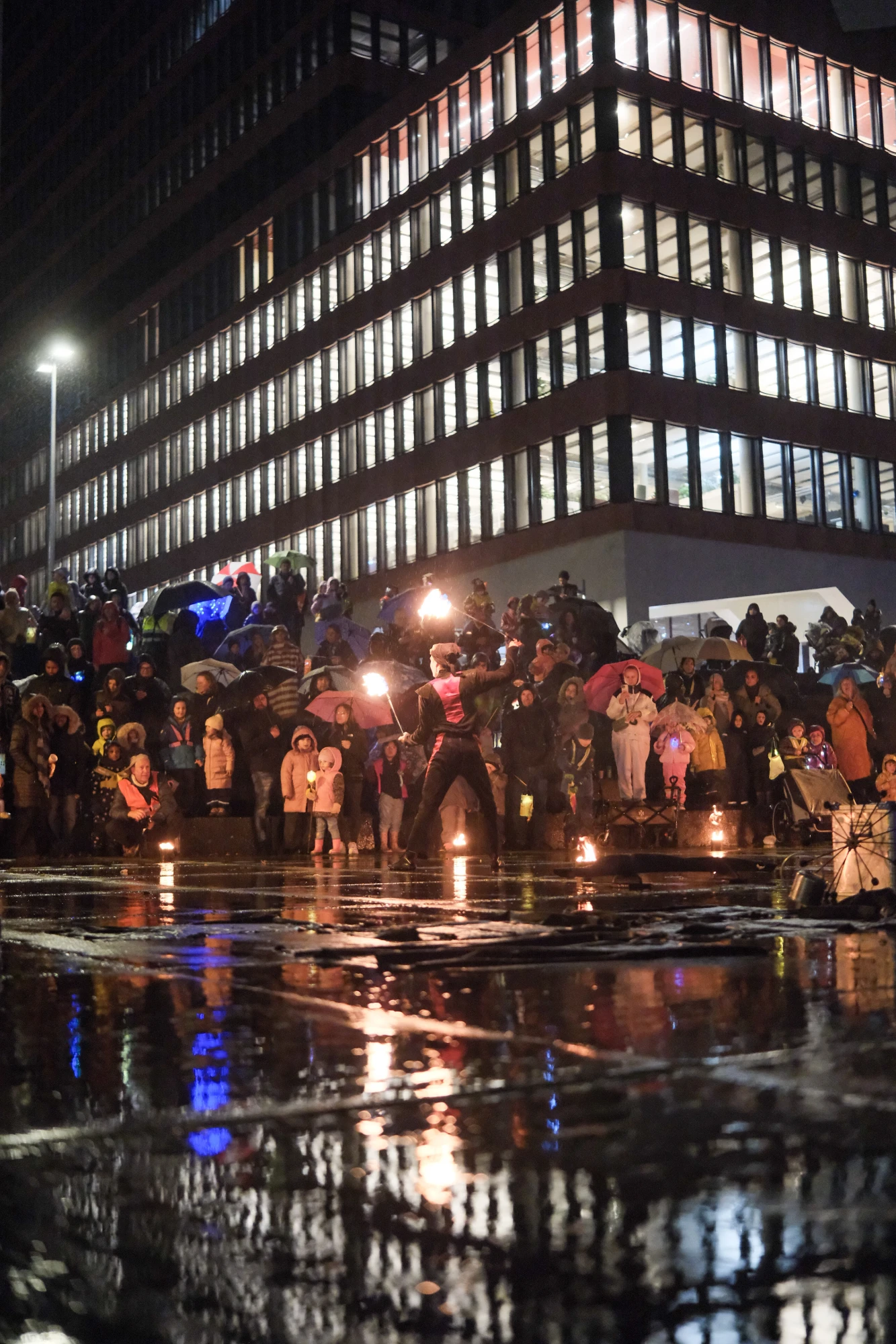 Feuerkünstler in Bewegung vor einer großen Menschenmenge bei Nacht, die mit Regenschirmen und warmen Kleidern zuschaut.