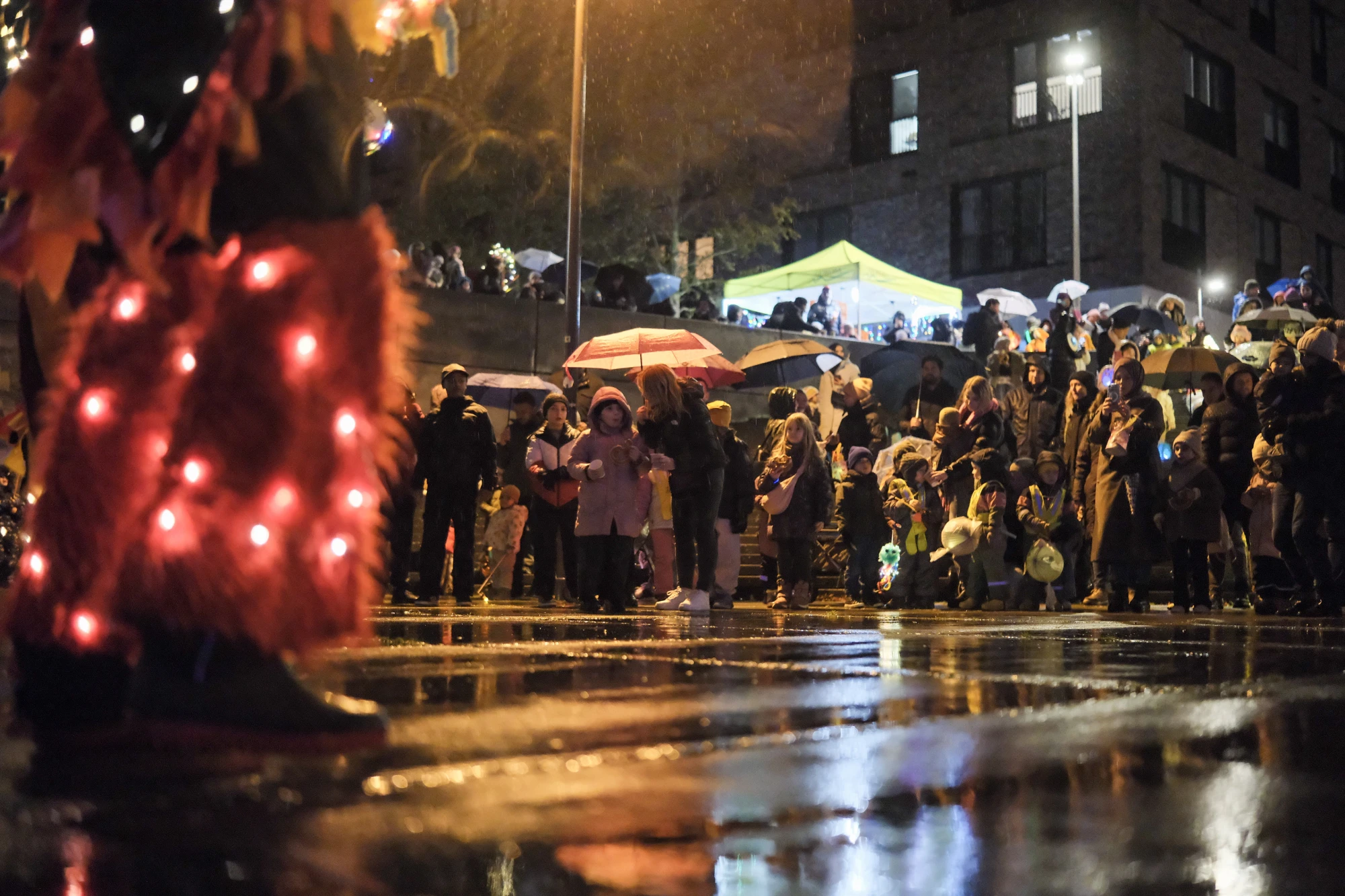 Menschen mit Regenschirmen und Laternen bei einer nächtlichen Veranstaltung auf nassem Straßenpflaster vor einem Gebäude